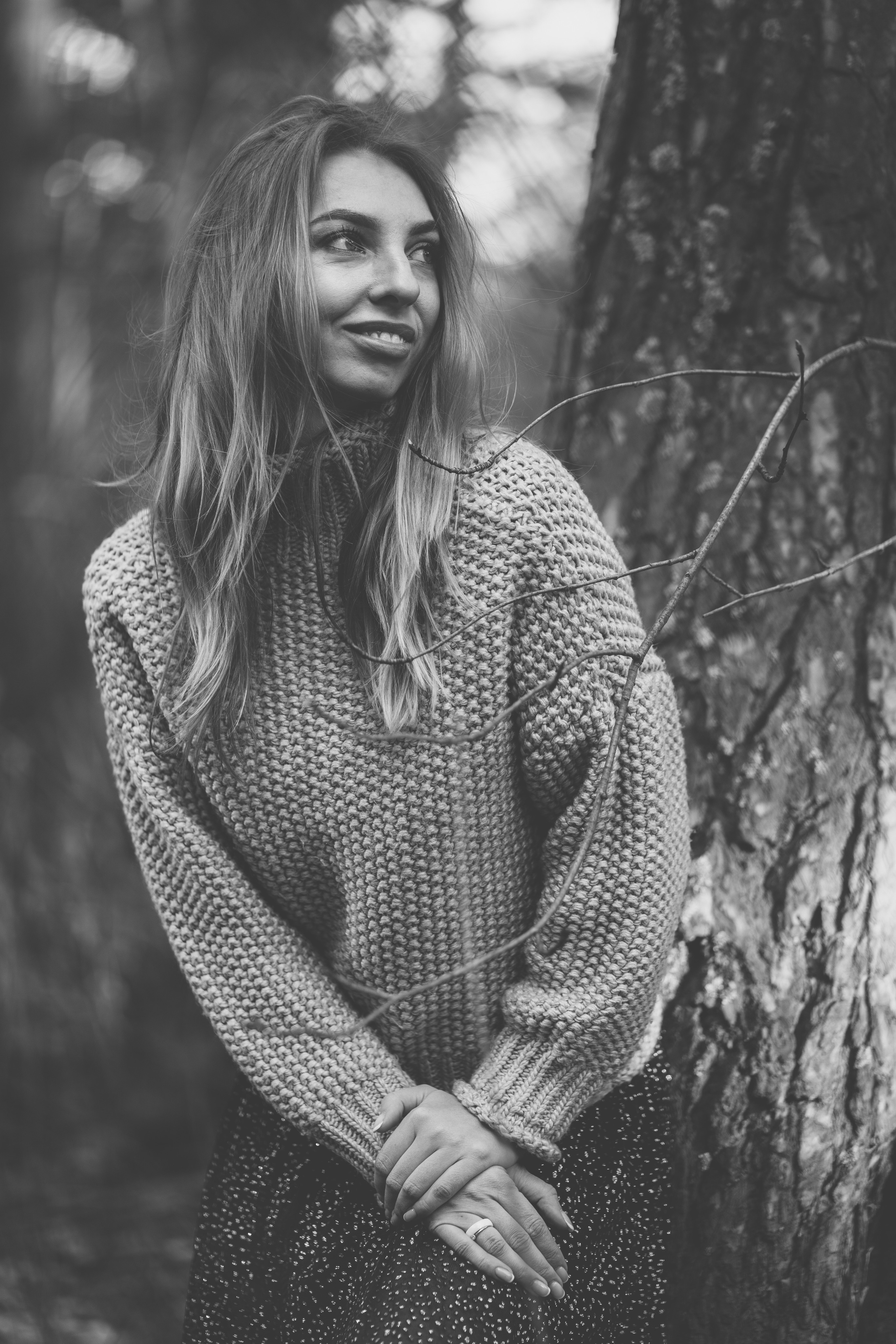 Девушка в вязанном свитере с горлом и легком платье, смотрит задорно не в камеру опираясь на дерево