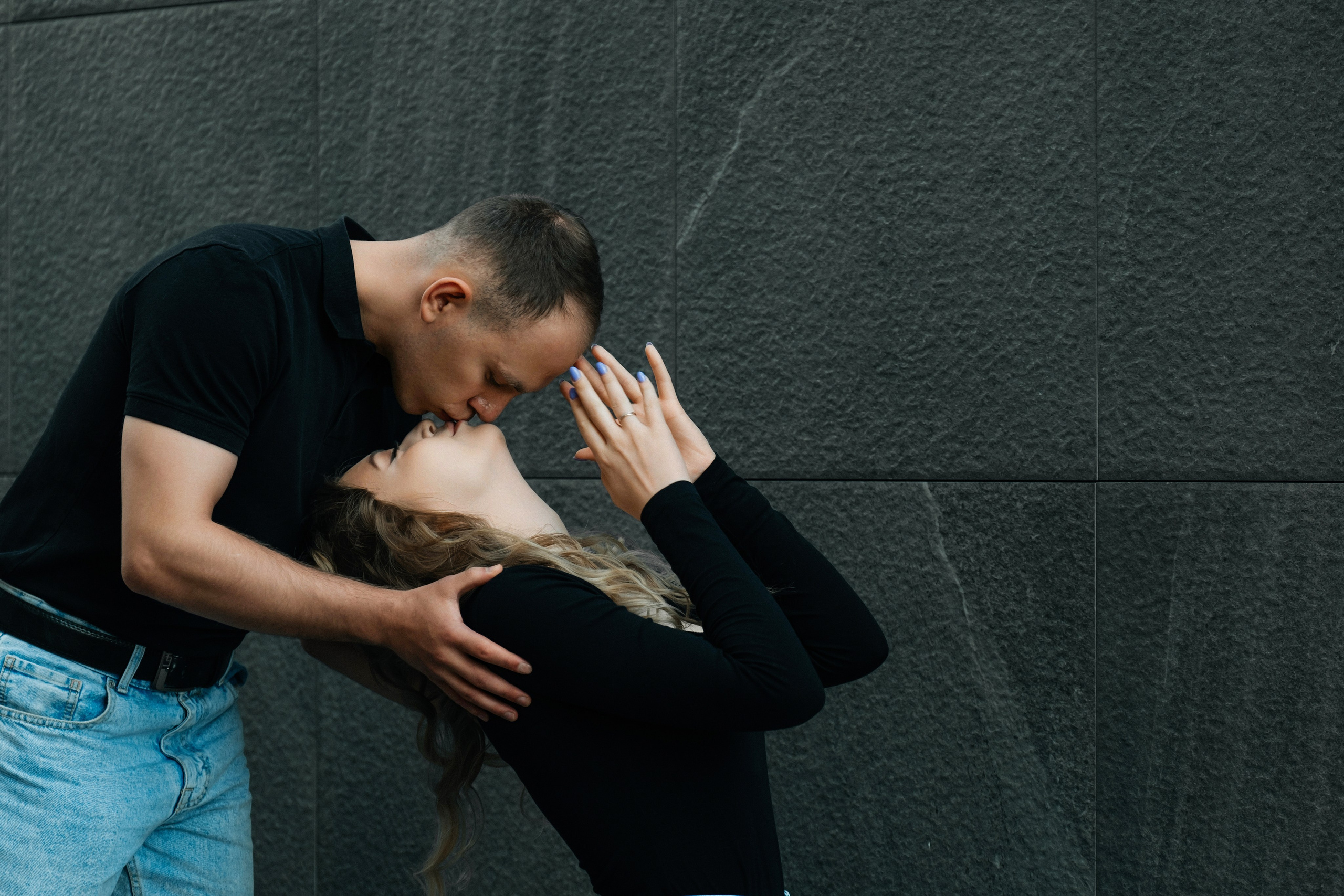 Молодая пара мужчина и девушка обнимаются у стены из серого камня в черных футболках и синих джинсах, лав стори для влюбленной пары на кануне свадьбы