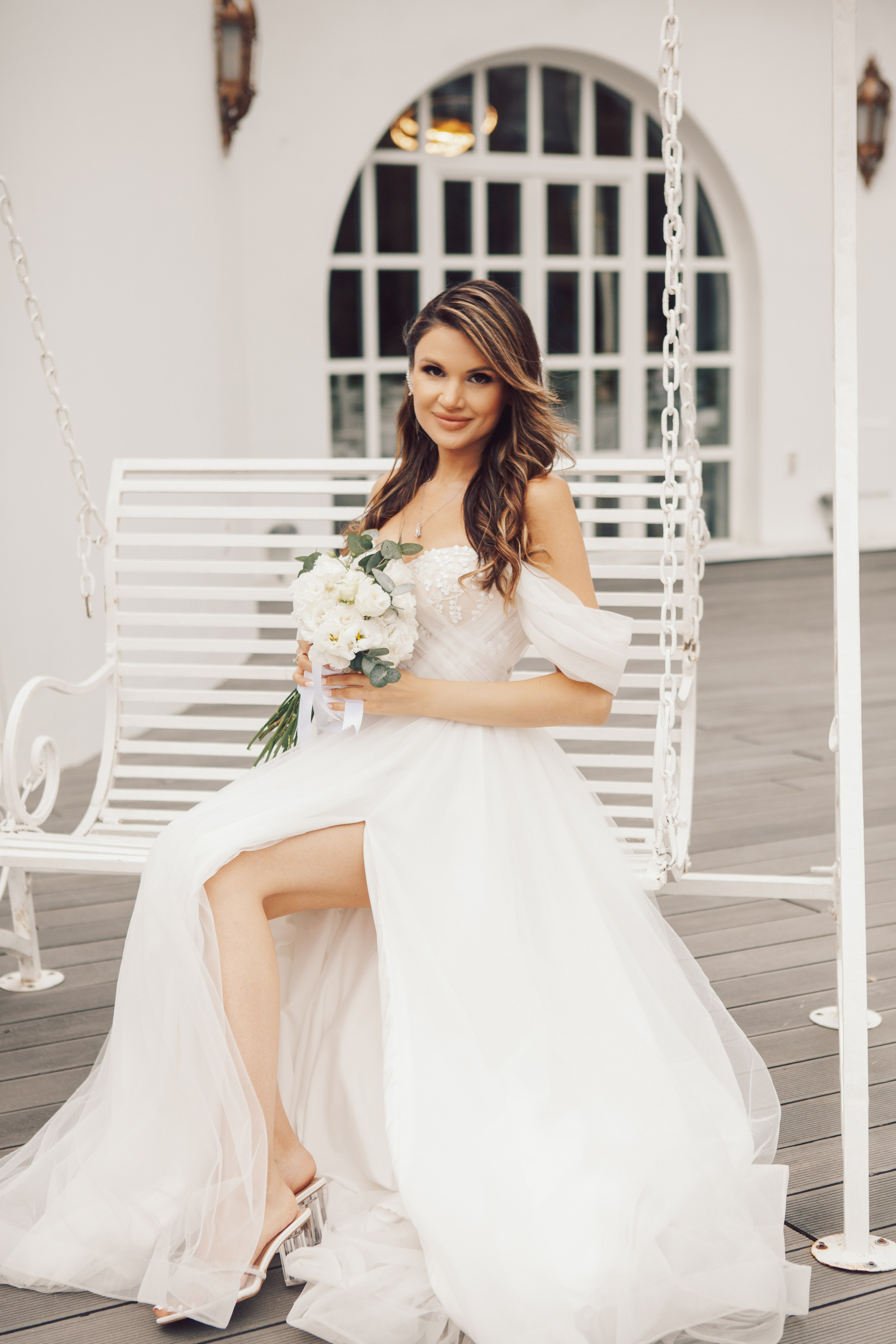 глубокий вырез на длинном платье у невесты, которая сидит на белых качелях