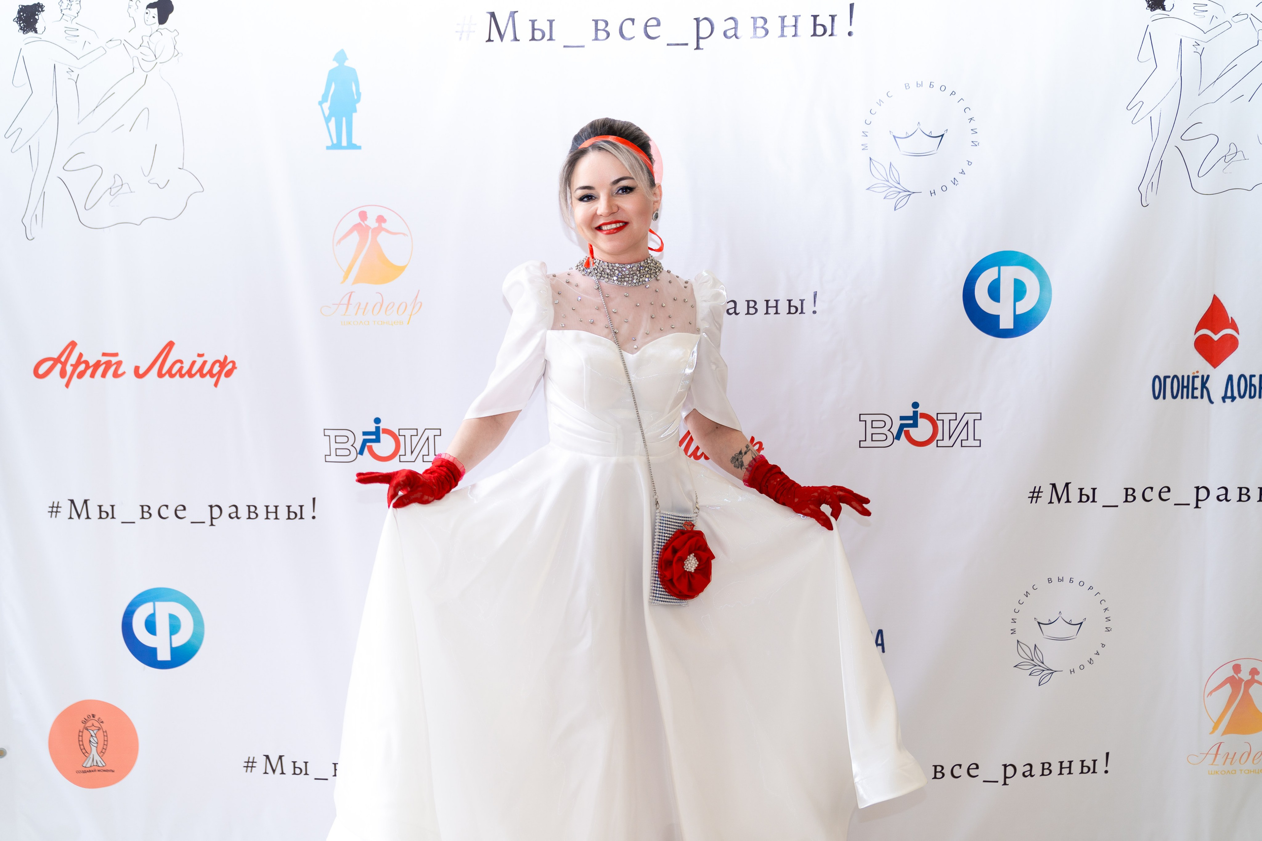 Инклюзивный бал «Мы все равны» — женщина в белом бальном платье с красными перчатками позирует на фоне баннера. Фотограф Мальцев Евгений, Санкт-Петербург.