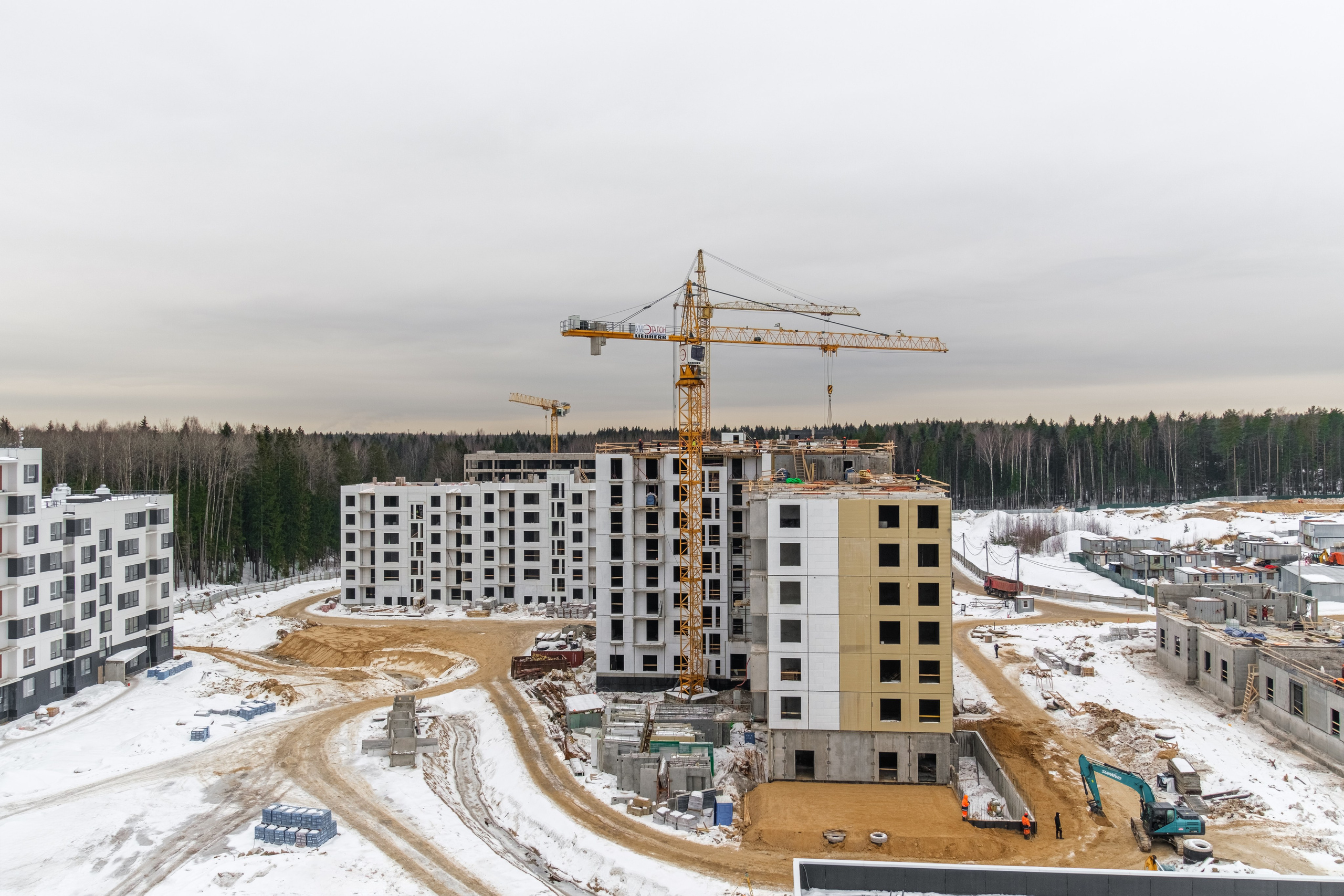 На изображении представлен процесс строительства жилых домов. Видно несколько зданий различной степени готовности: некоторые уже построены и облицованы, другие находятся на разных стадиях возведения. Также присутствует строительная техника, включая краны. Строительная площадка покрыта снегом, что указывает на зимний период.