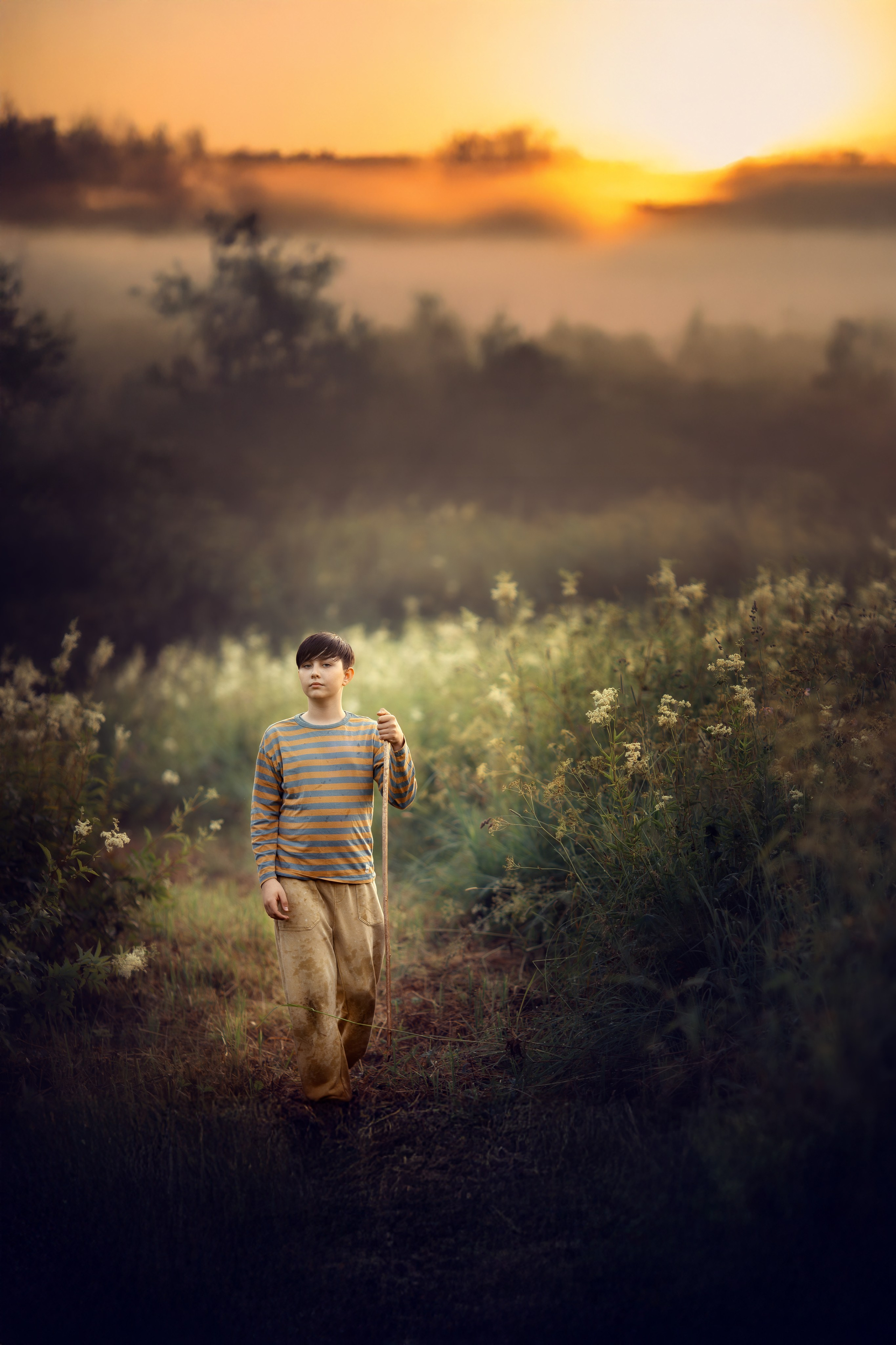 Мальчик в полосатой кофте идёт по заросшей тропинке в туманный вечер, на фоне — закатное небо и мягкий свет.
