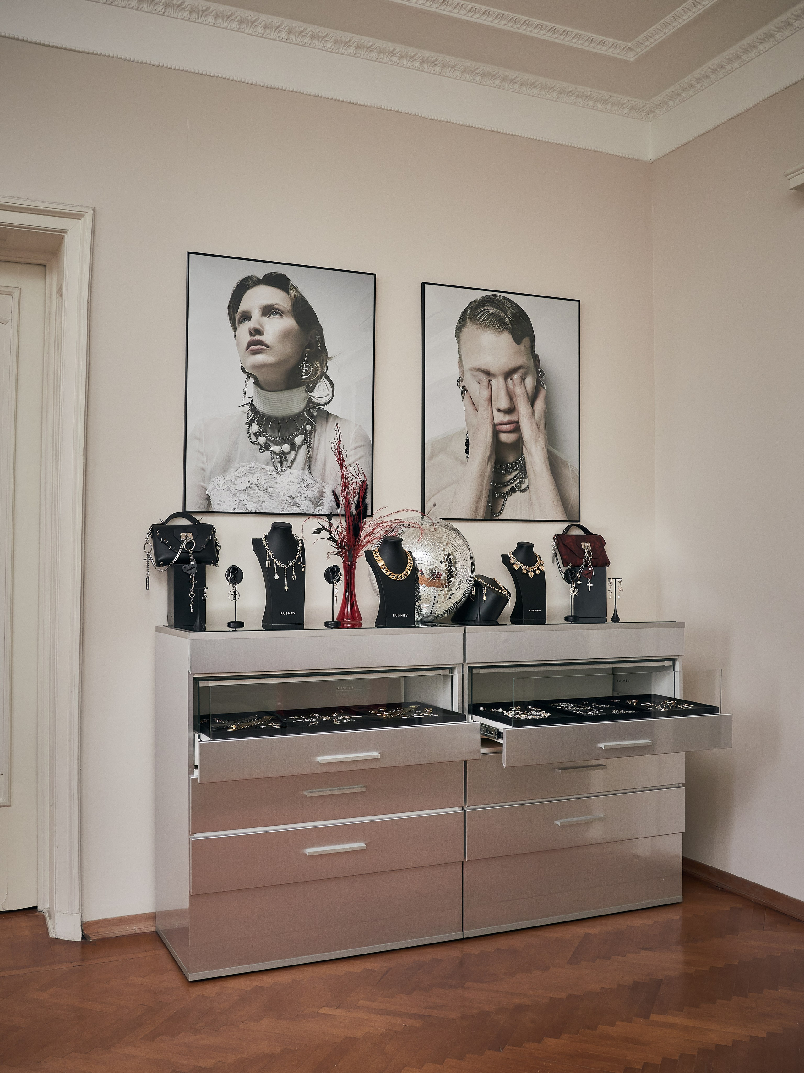 В стильном шоуруме есть комод с открытыми ящиками, в которых хранятся драгоценности. Выше висят два больших черно-белых модных портрета, на которых изображена модель с аксессуарами. На верху комода находятся различные ожерелья, браслеты и декоративная ваза с красными ветвями.