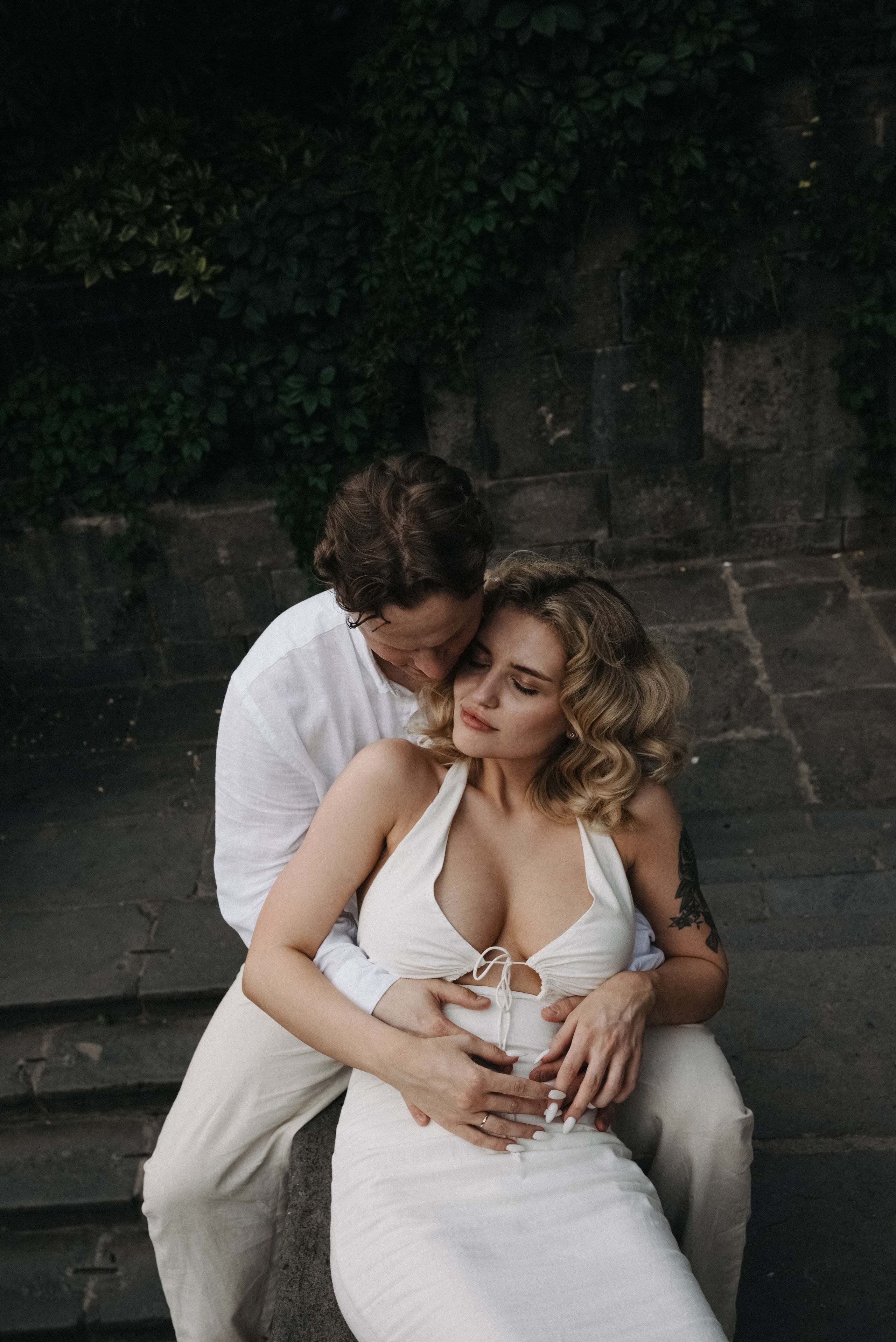 Невеста в белом платье спиной оперлась на жениха в белом костюме и смотрит на него