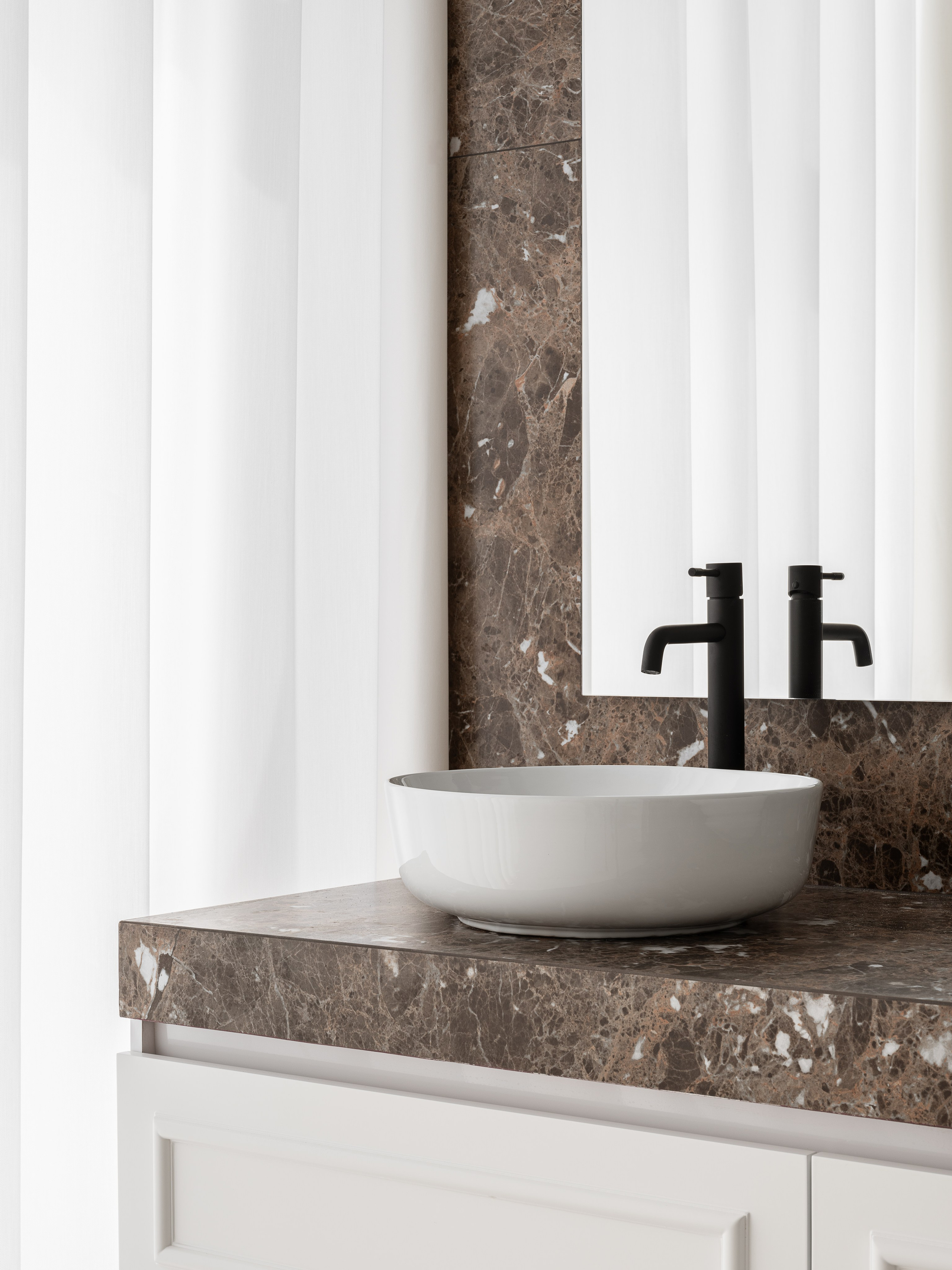 Современная деталь ванной комнаты с белой круглой раковиной-чашей, коричневой мраморной столешницей и черным смесителем на фоне белых штор, созданная дизайнером Василиссой Бачевой, стилистом Натальей Якушевой и сфотографированная Сергеем Денисенко.