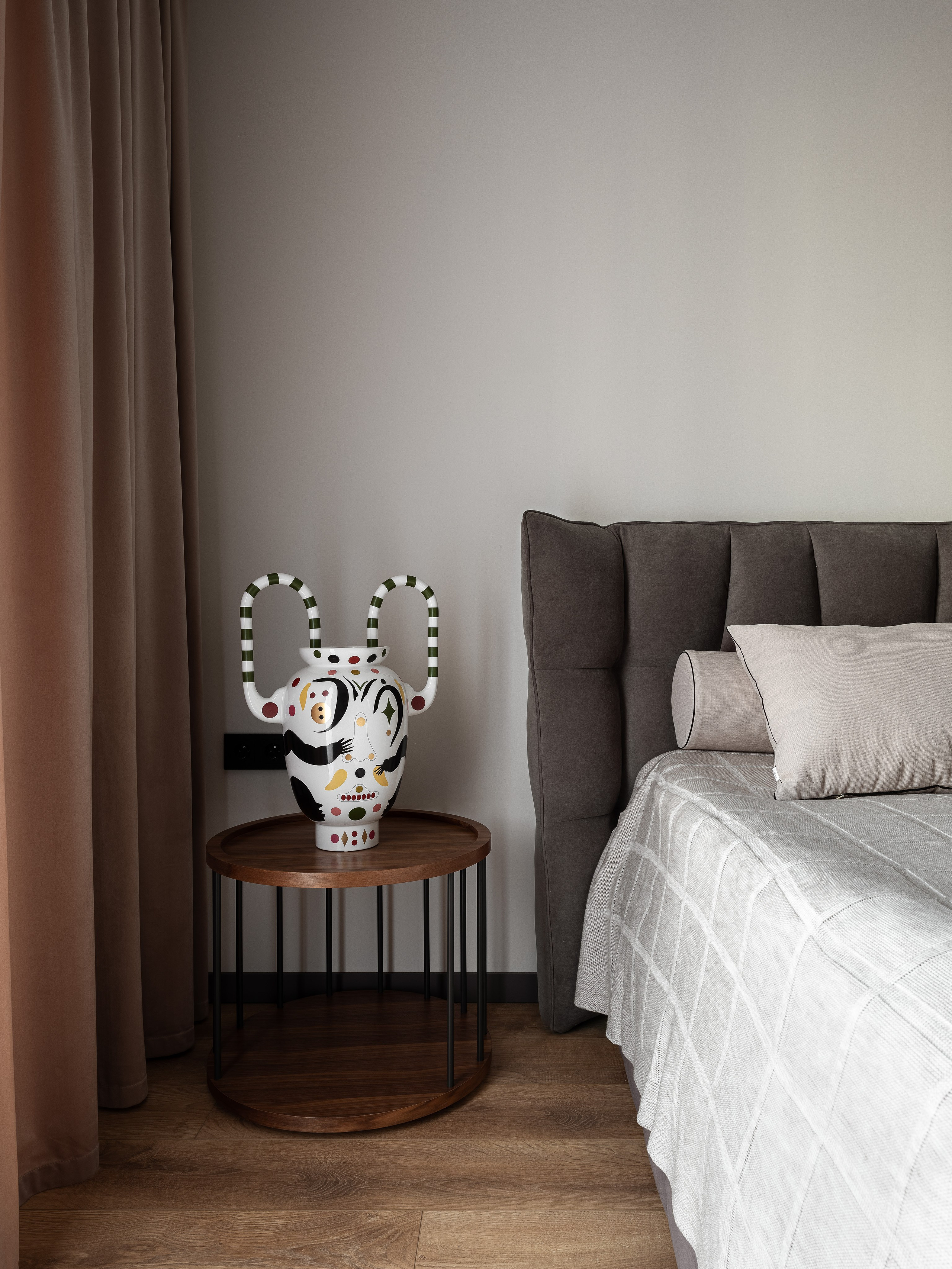 Современная спальня с серым изголовьем кровати, прикроватным круглым столиком и авторской вазой с ярким орнаментом, дизайнер Екатерина Мищенко, фотограф Сергей Денисенко, гармоничное сочетание акцентных деталей и спокойных оттенков.