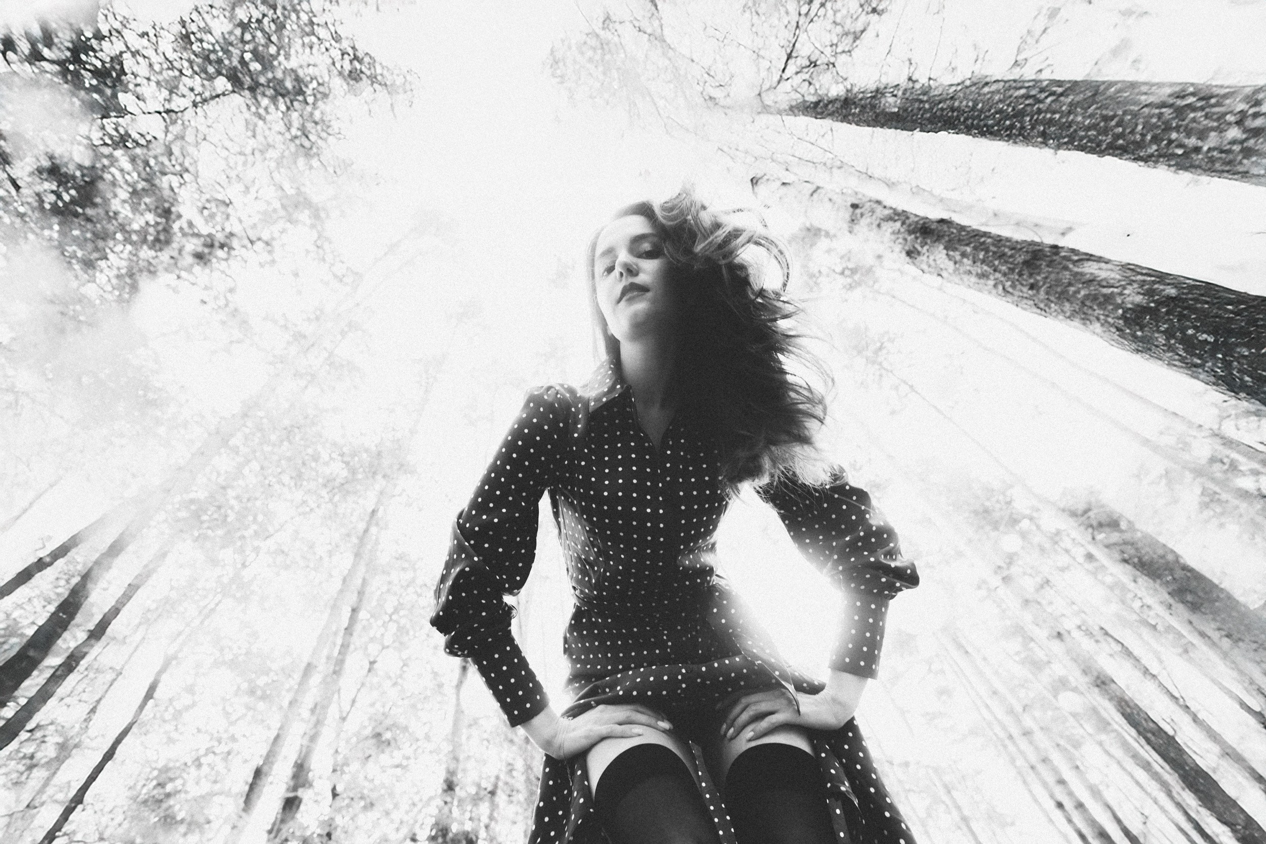 Чёрно-белый портрет в лесу: женщина в платье в горошек и чулках, снятая снизу на фоне высоких деревьев