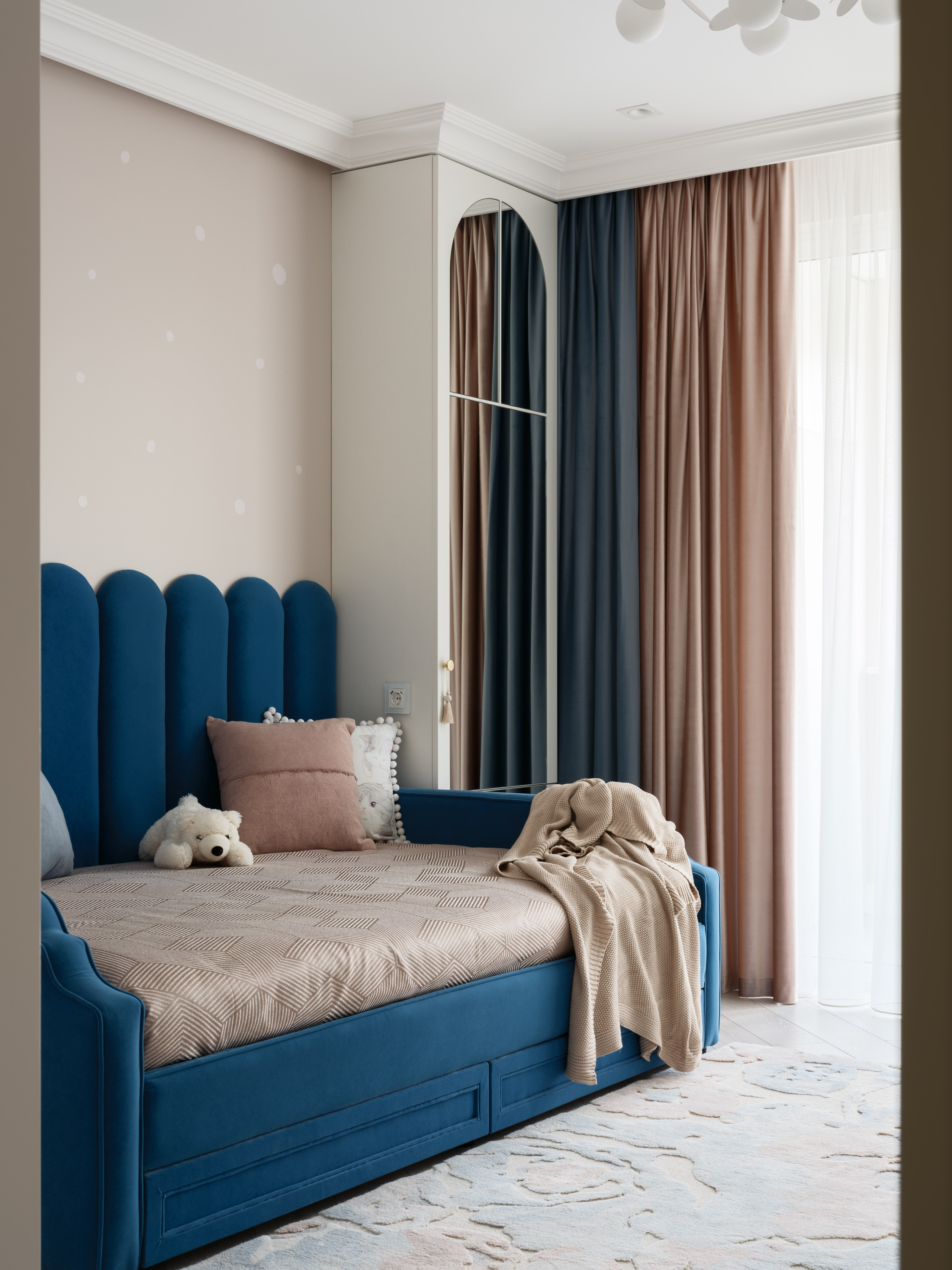 Уютная детская с синим диваном-кроватью, мягкими подушками и игрушкой. На стене спокойный декор, шторы в бежево-розовых тонах. Проект Евгении Ермошиной, фотограф Сергей Денисенко.
