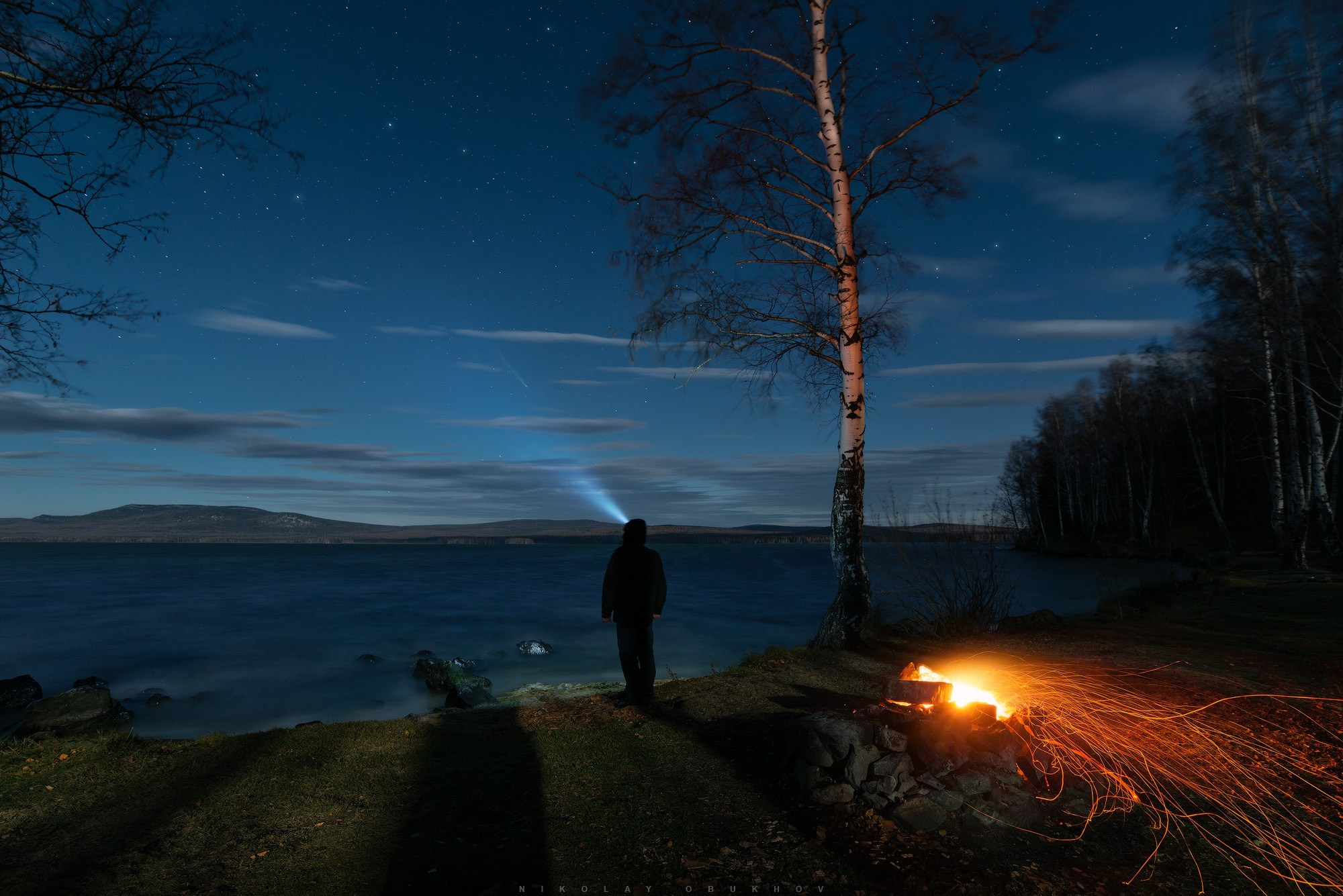 Человек наблюдает комету C/2023 A3 (Tsuchinshan-ATLAS) на берегу озера в октябре 2024 года.