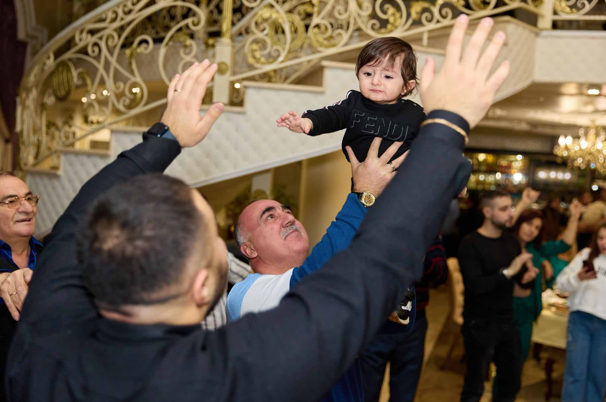 Дедушка поднимает именинника над головой, гости рядом