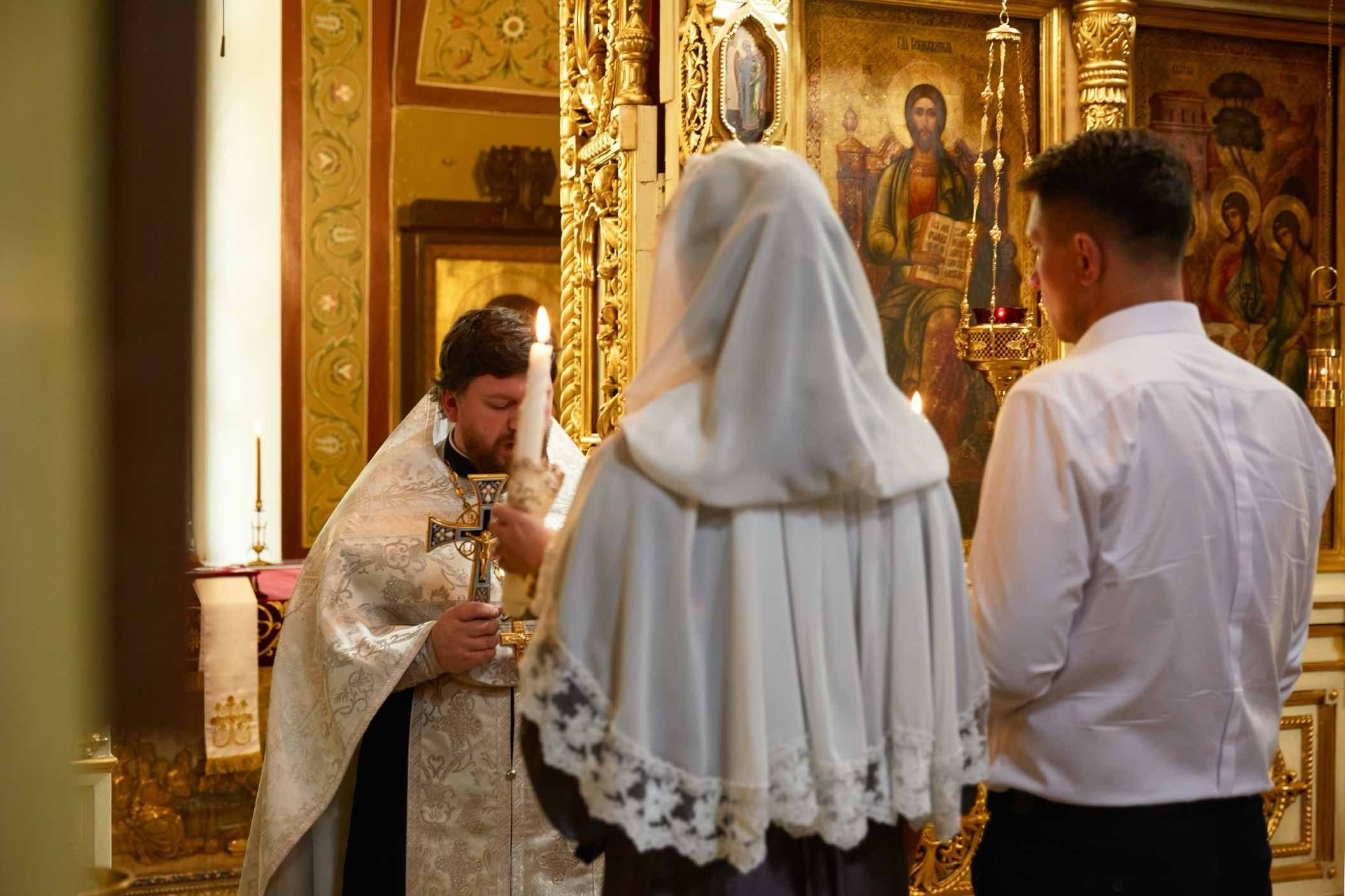 Священник с крестом перед венчающейся парой, кадр снят из-за спины, невеста со свечой под фатой в храме города Лобня