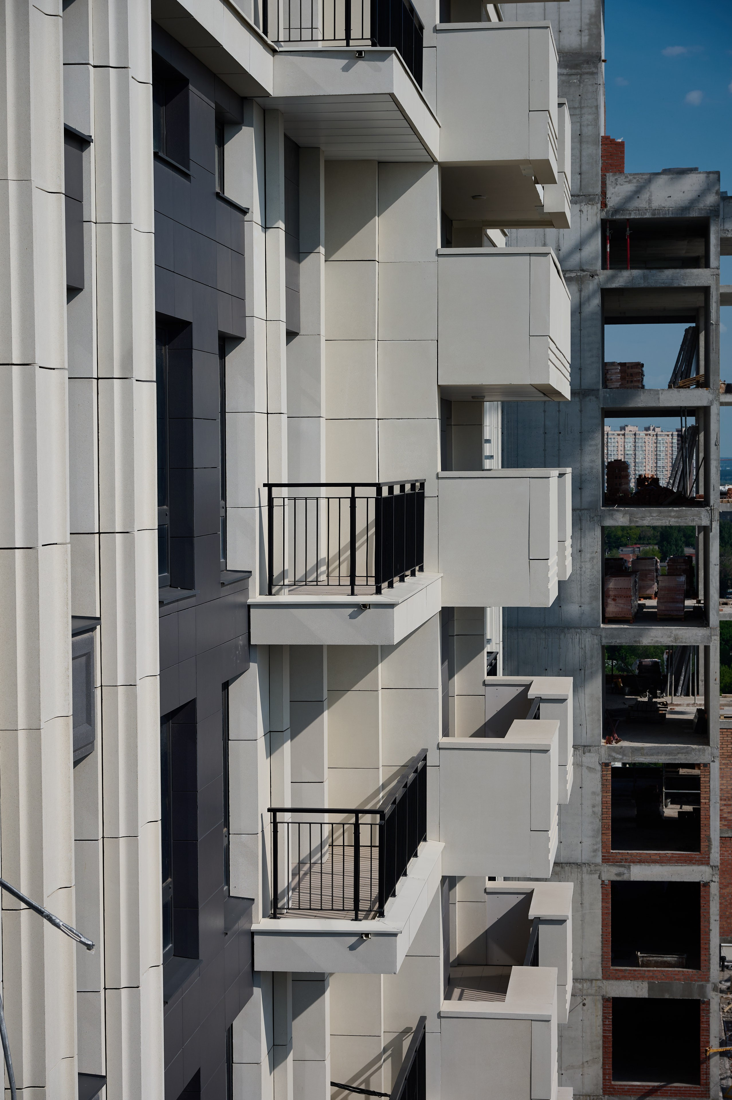 Фасад жилого дома с белой отделкой и узкими балконами; справа видны фрагменты соседнего здания на стадии строительства