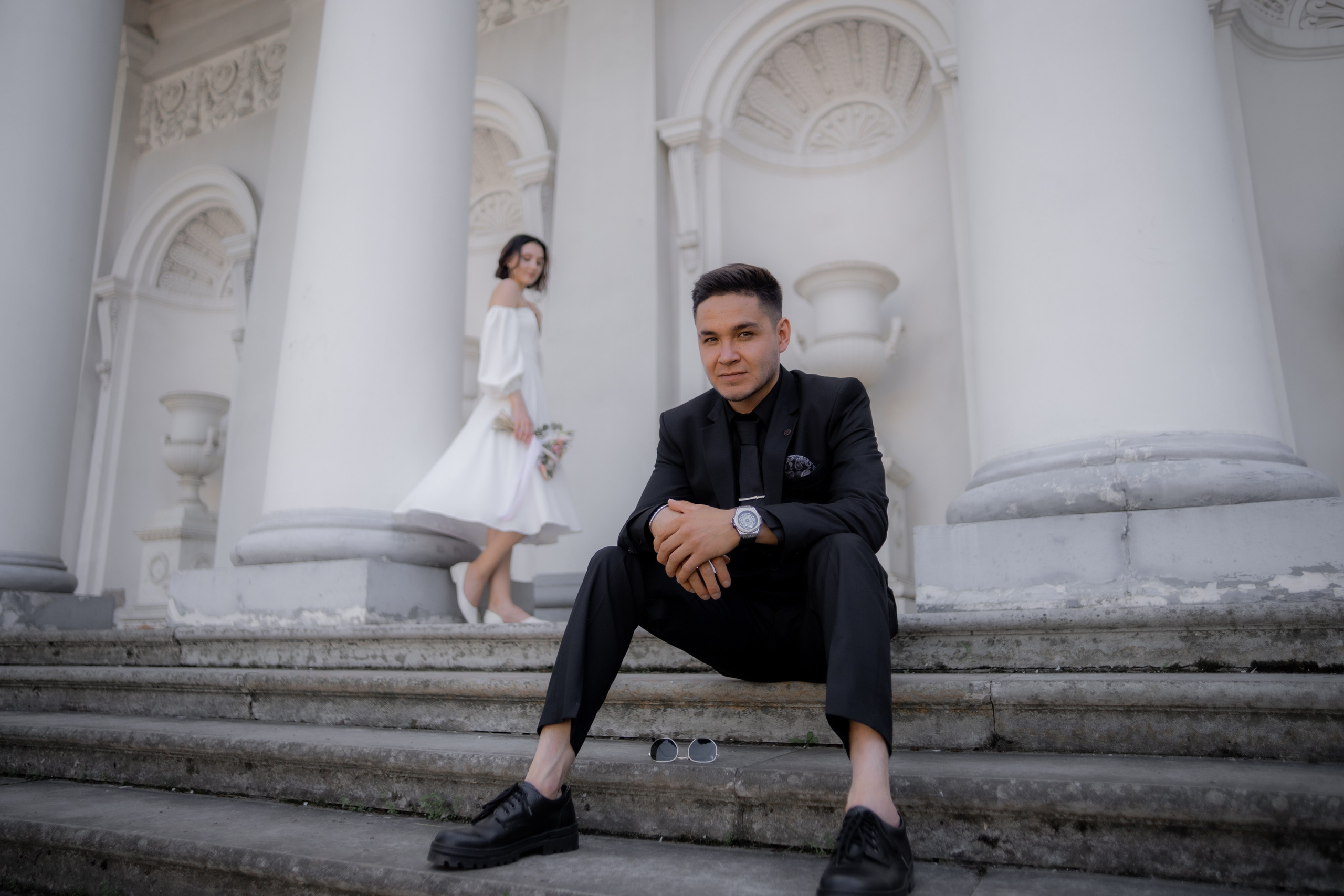 передний план - жених в черном костюме, задний план - невеста в платье с открытыми плечами  на елагином острове