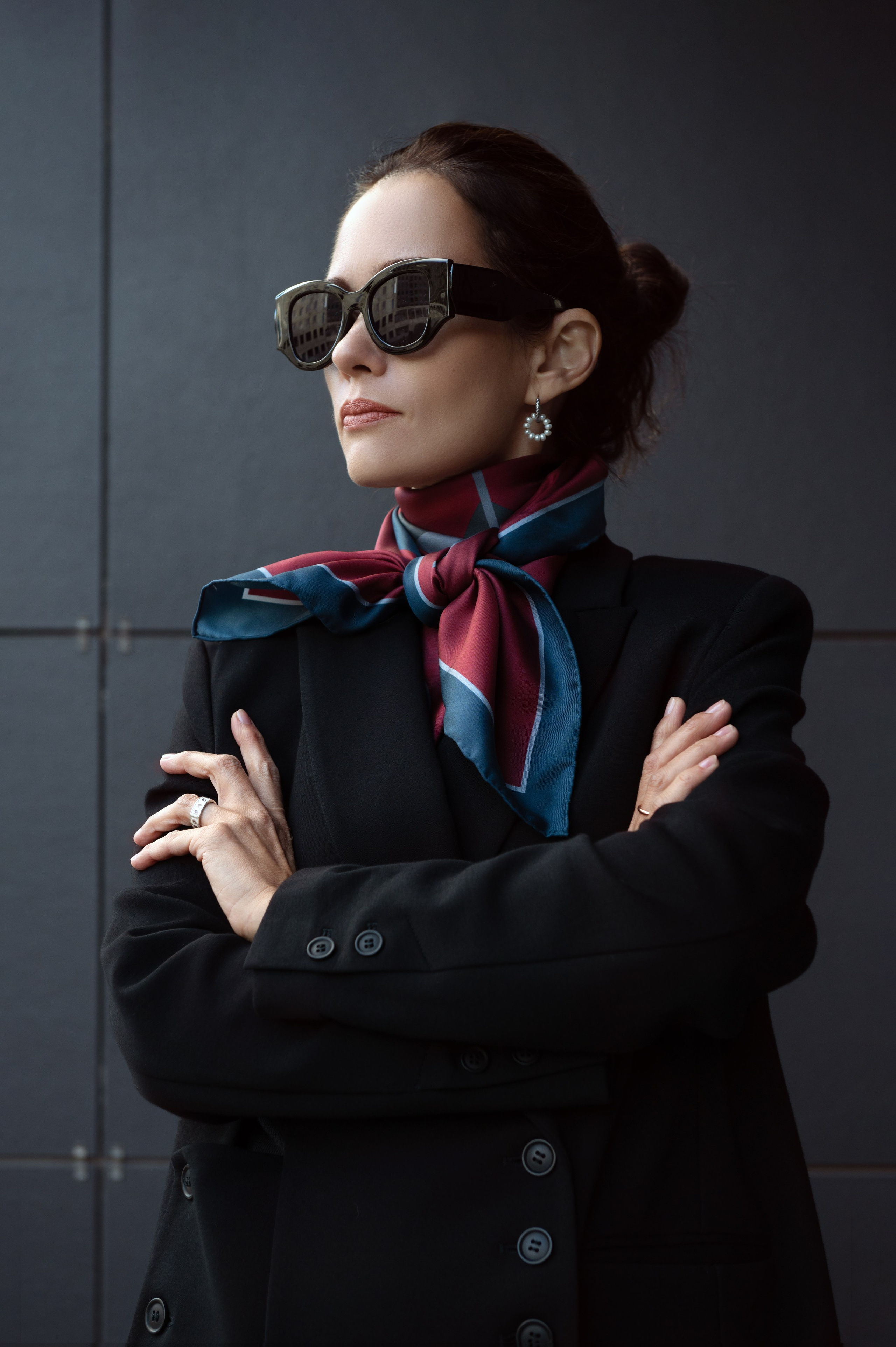 Эффектный портрет девушки с естественным свете. Девушка повернута в три четверти к камере, руки мягко скрещены на груди, съемка чуть снизу, чтобы добавить властности образу. Свет падает слева сверху, красиво подчеркивая фактуру одежды и скулы модели. Уличный портрет