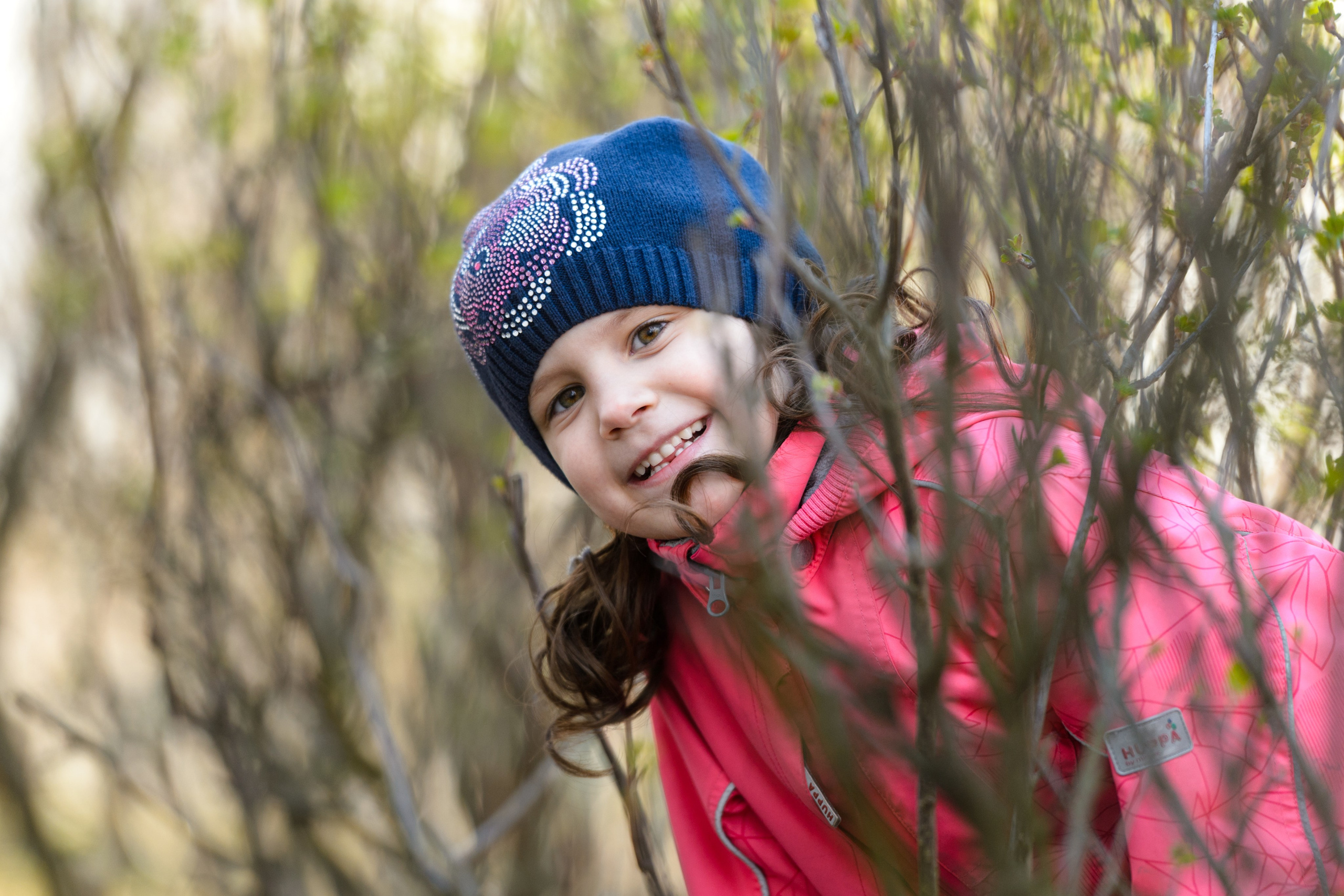 Девочка в синей шапке и розовой куртке улыбается на улице, выглядывая из-за веток.