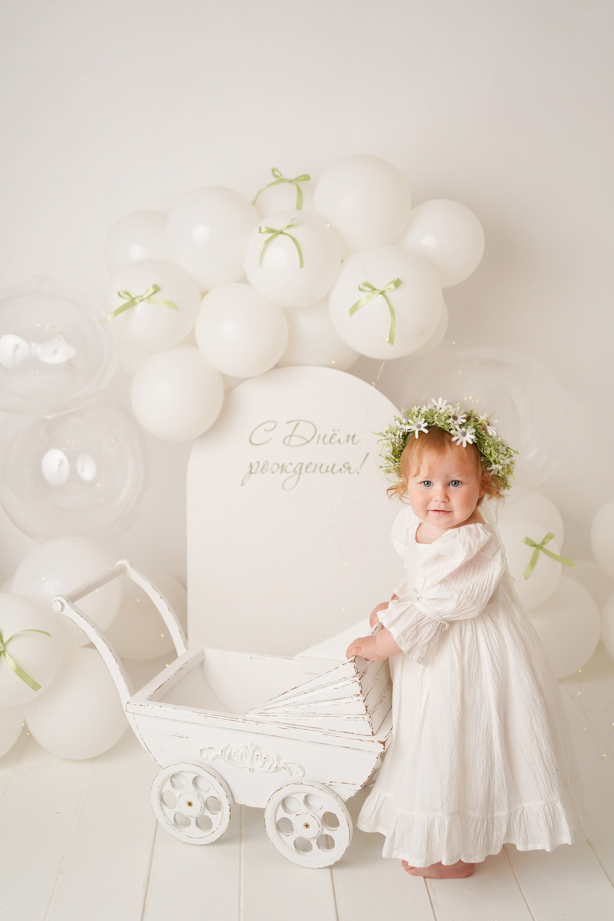 Малышка в белом платье и веночке из цветов стоит рядом с колясочкой.