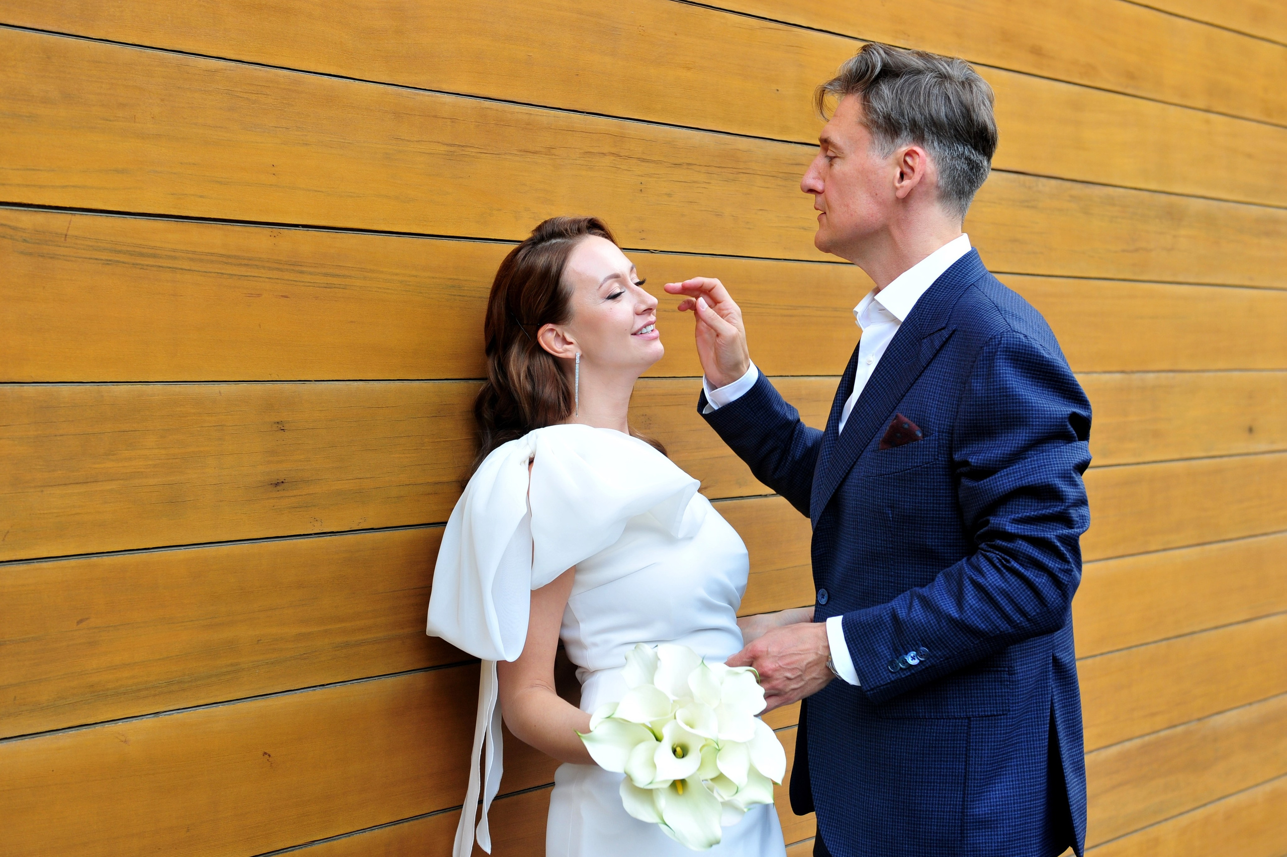 Свадьба в ЗАГСе Барвиха Luxury Village летом, жених и невеста стоят у деревянной стены здания и смотрят друг на друга