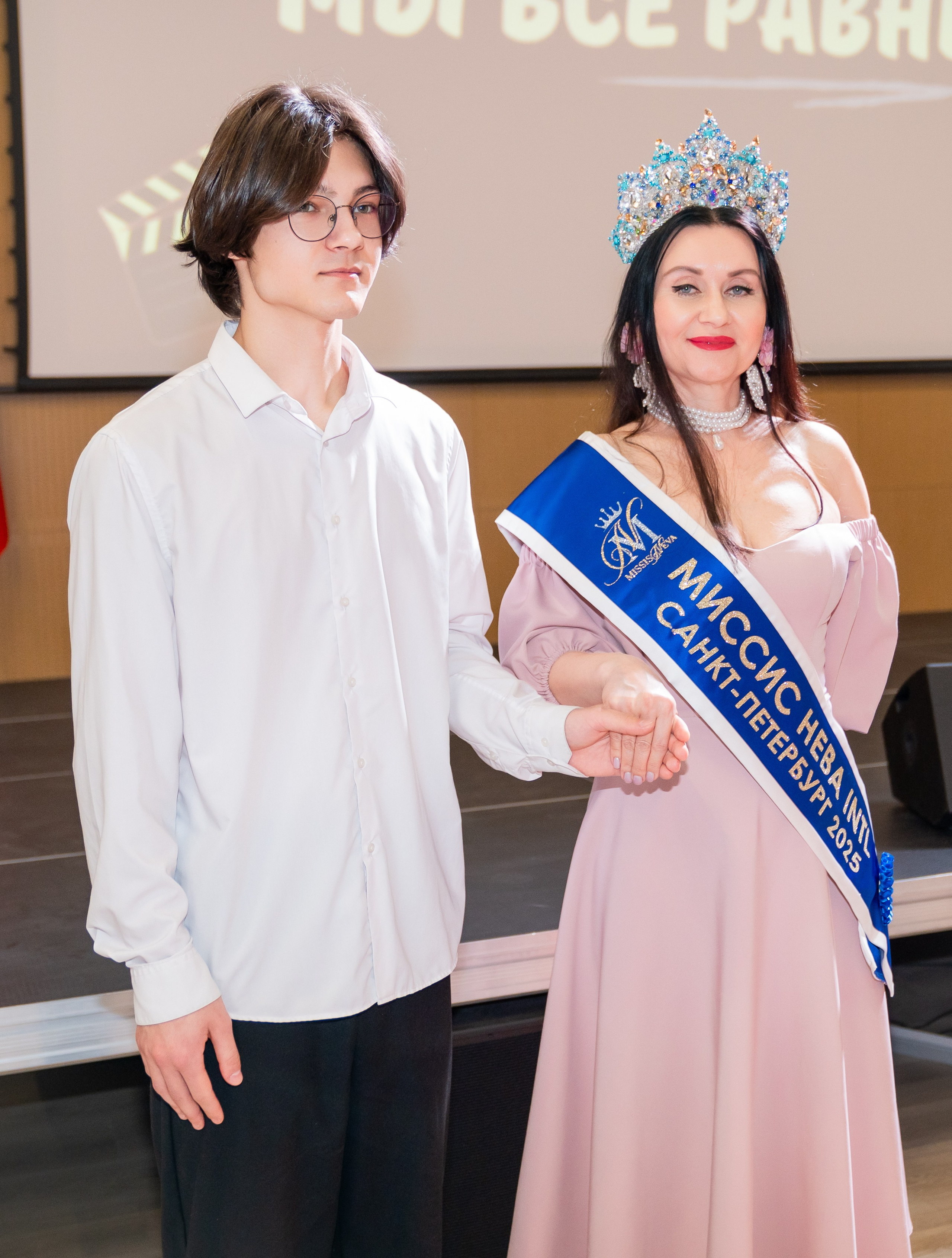 Молодой человек в белой рубашке держит за руку Мисс Санкт-Петербург International 2025 в розовом платье и короне. Инклюзивный бал «Мы все равны». Фотограф Мальцев Евгений.