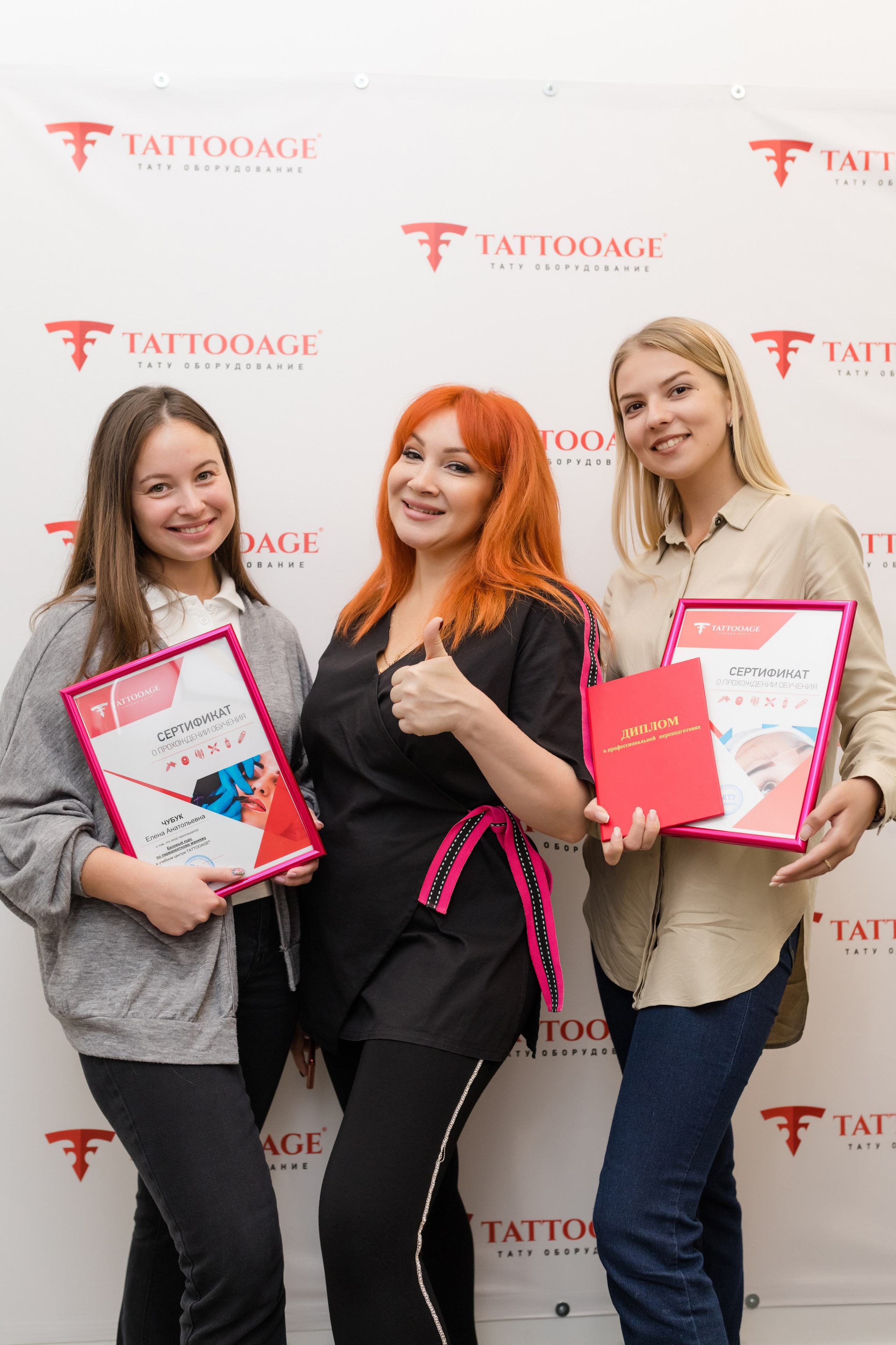 Две ученицей и преподаватель стоят на фоне баннера с дипломами о окончании курсов татуажа