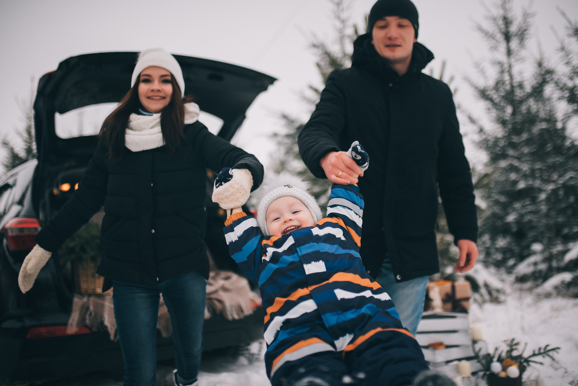 Зимняя семейная фотосессия у автомобиля. Папа с мамой качают ребенка