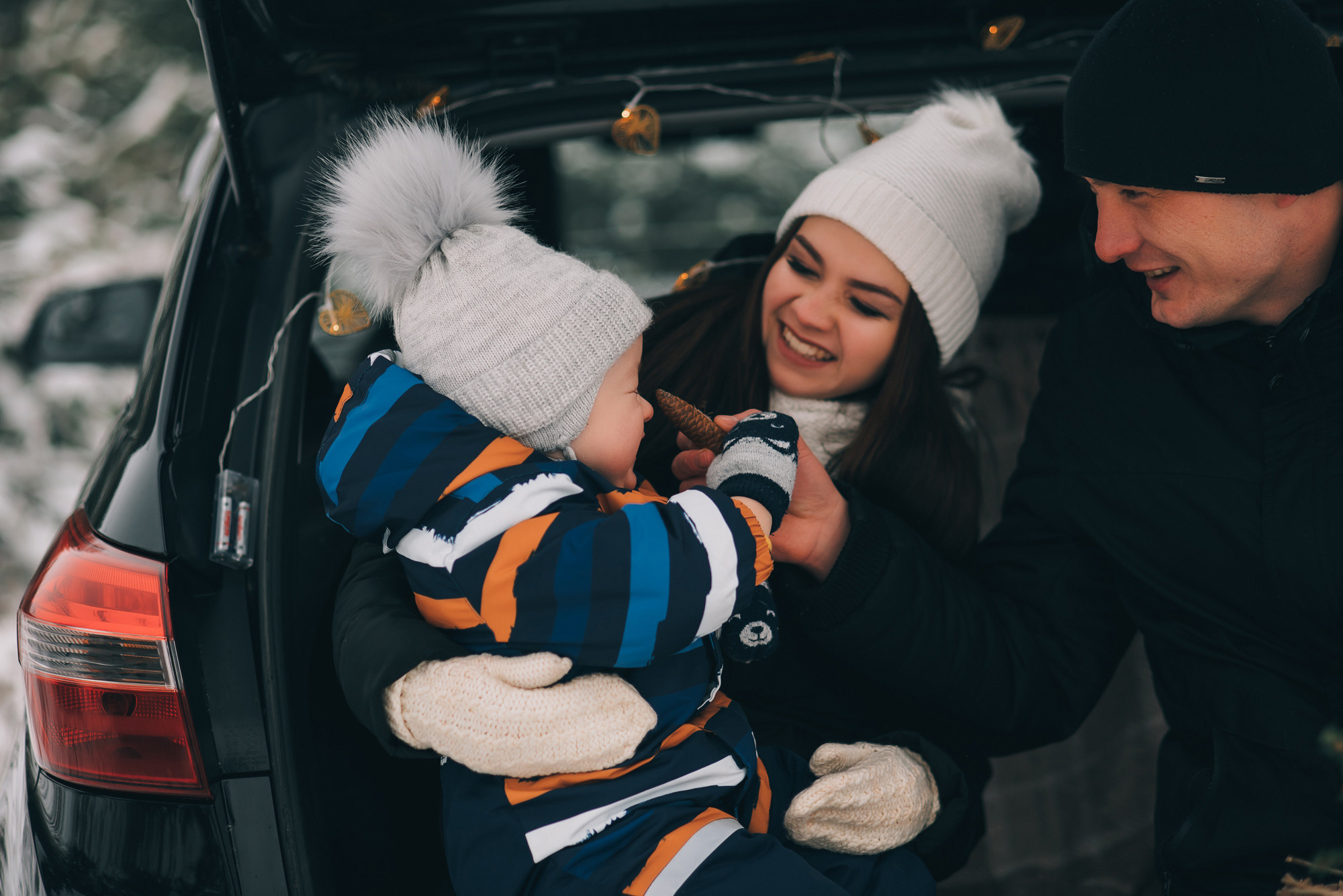 Зимняя семейная фотосессия у автомобиля. Папа с мамой смотрят на ребенка с улыбкой