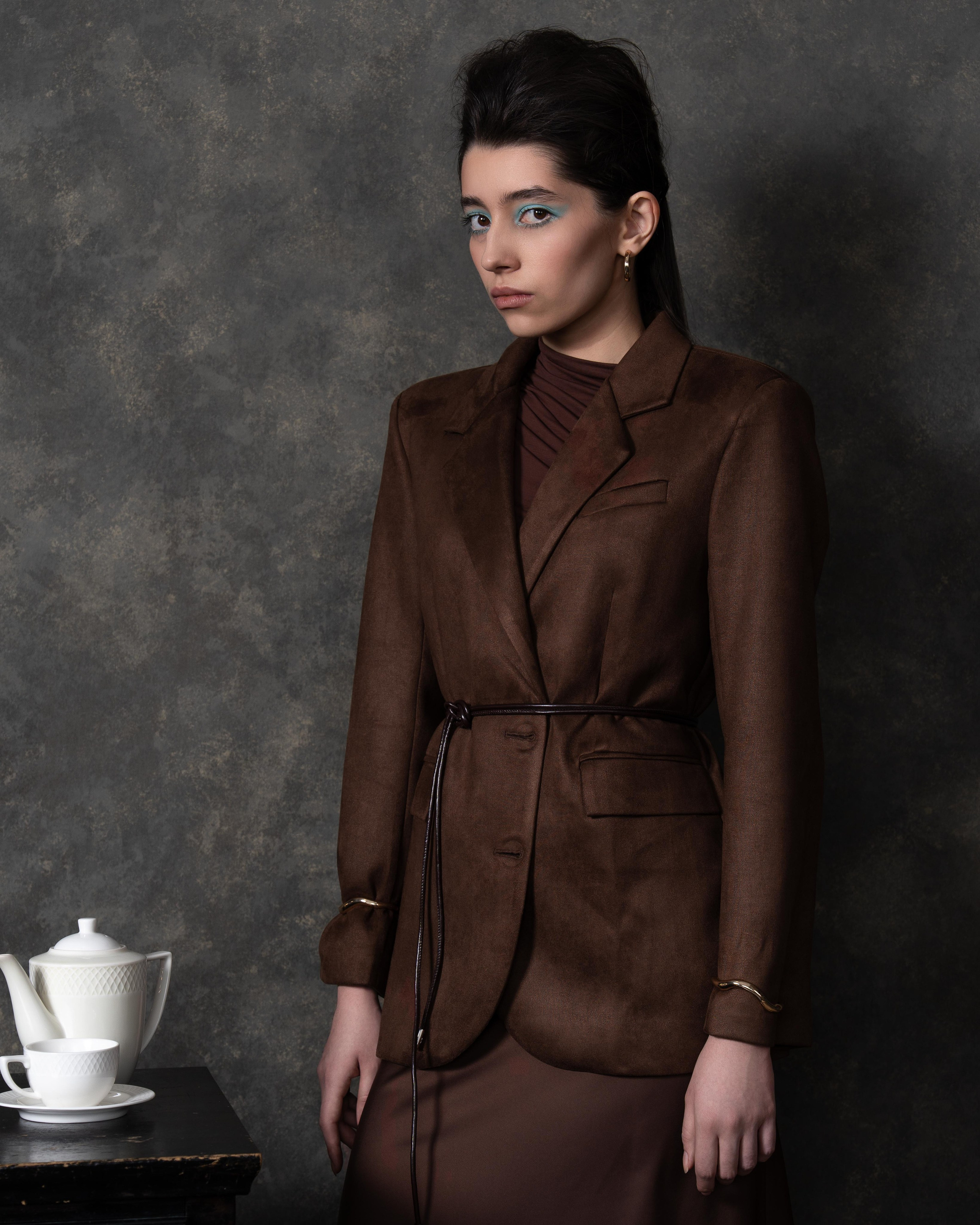 На фотографии изображена женщина, одетая в коричневый костюм с поясом. Она стоит на фоне серой стены, которая создает контраст с ее одеждой. На столе рядом с ней стоит белый чайный сервиз. Женщина смотрит прямо в камеру, ее взгляд серьезен и сосредоточен.