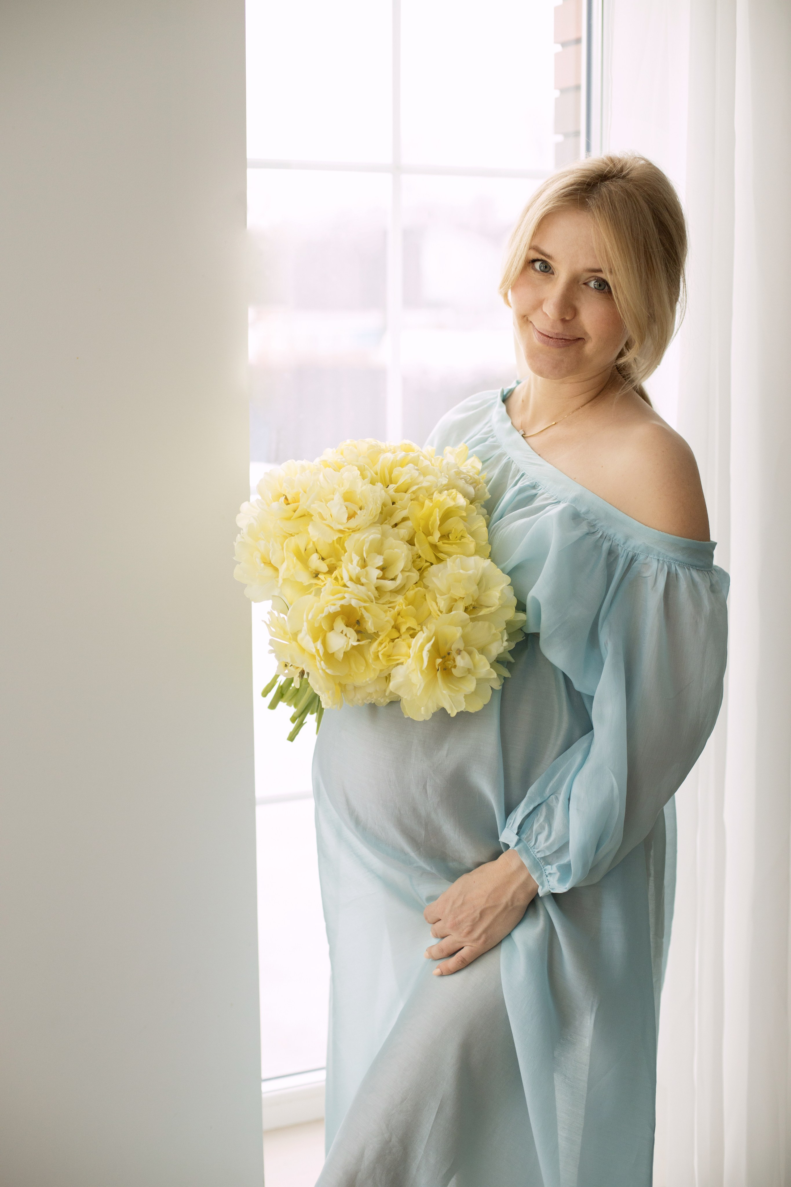 Беременная женщина в  голубом платье с букетом желтых пионов стоит у большого окна.