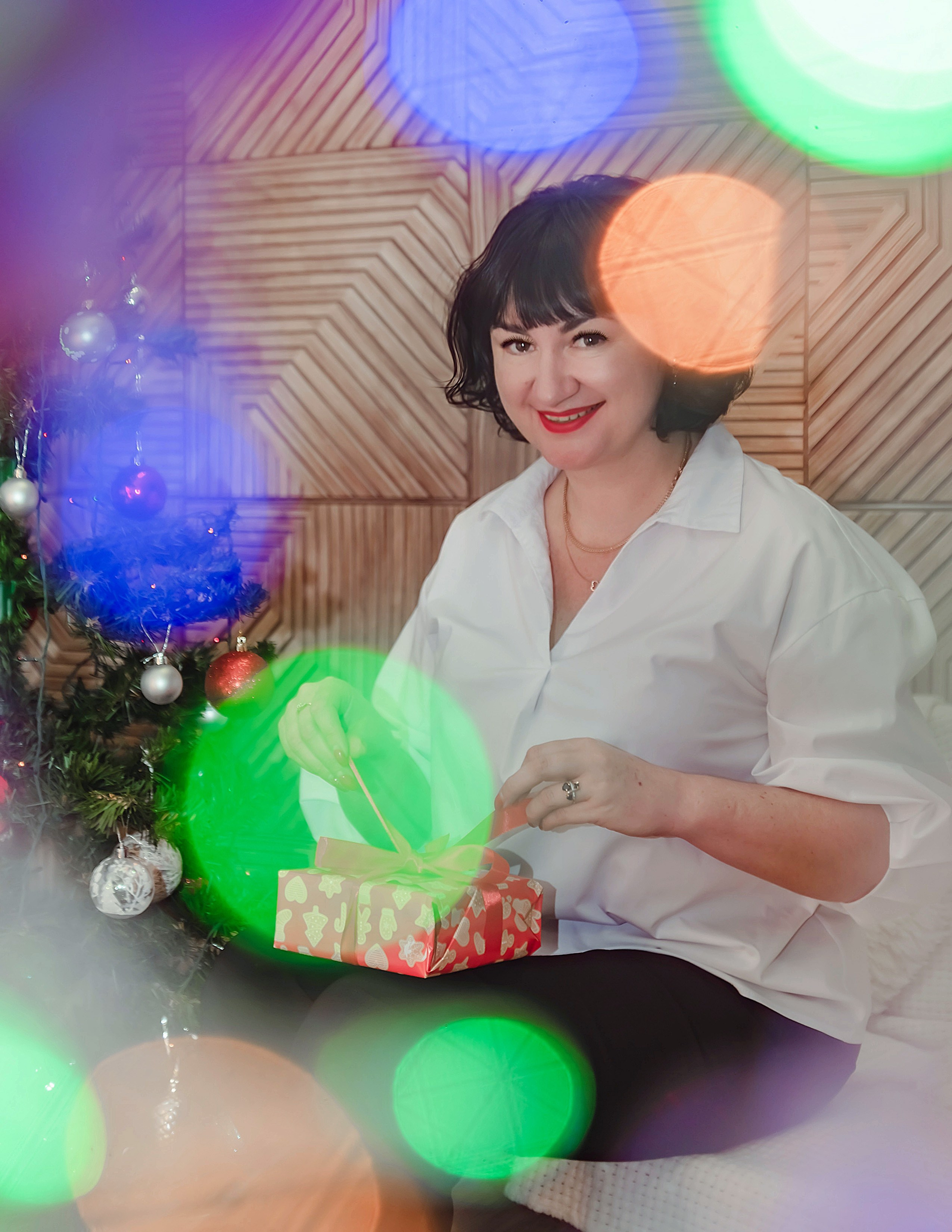 На фото девушка с подарком и огоньками на переднем плане.