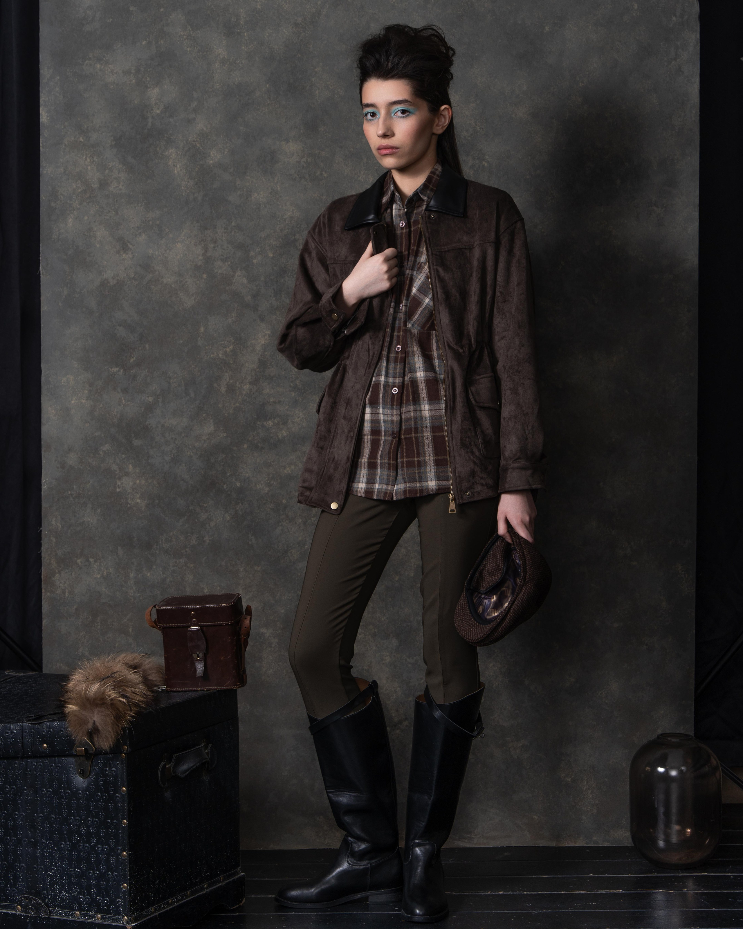 На фотографии изображена женщина, стоящая на фоне серой стены. Она одета в коричневую куртку с меховым воротником и клетчатую рубашку под ней. На ней также зеленые брюки и черные сапоги. В одной руке она держит коричневую шляпу, а в другой — коричневую сумку. Рядом с ней стоит черный чемодан с меховой отделкой и черная ваза. Женщина смотрит прямо в камеру, ее взгляд сосредоточен и уверенный.