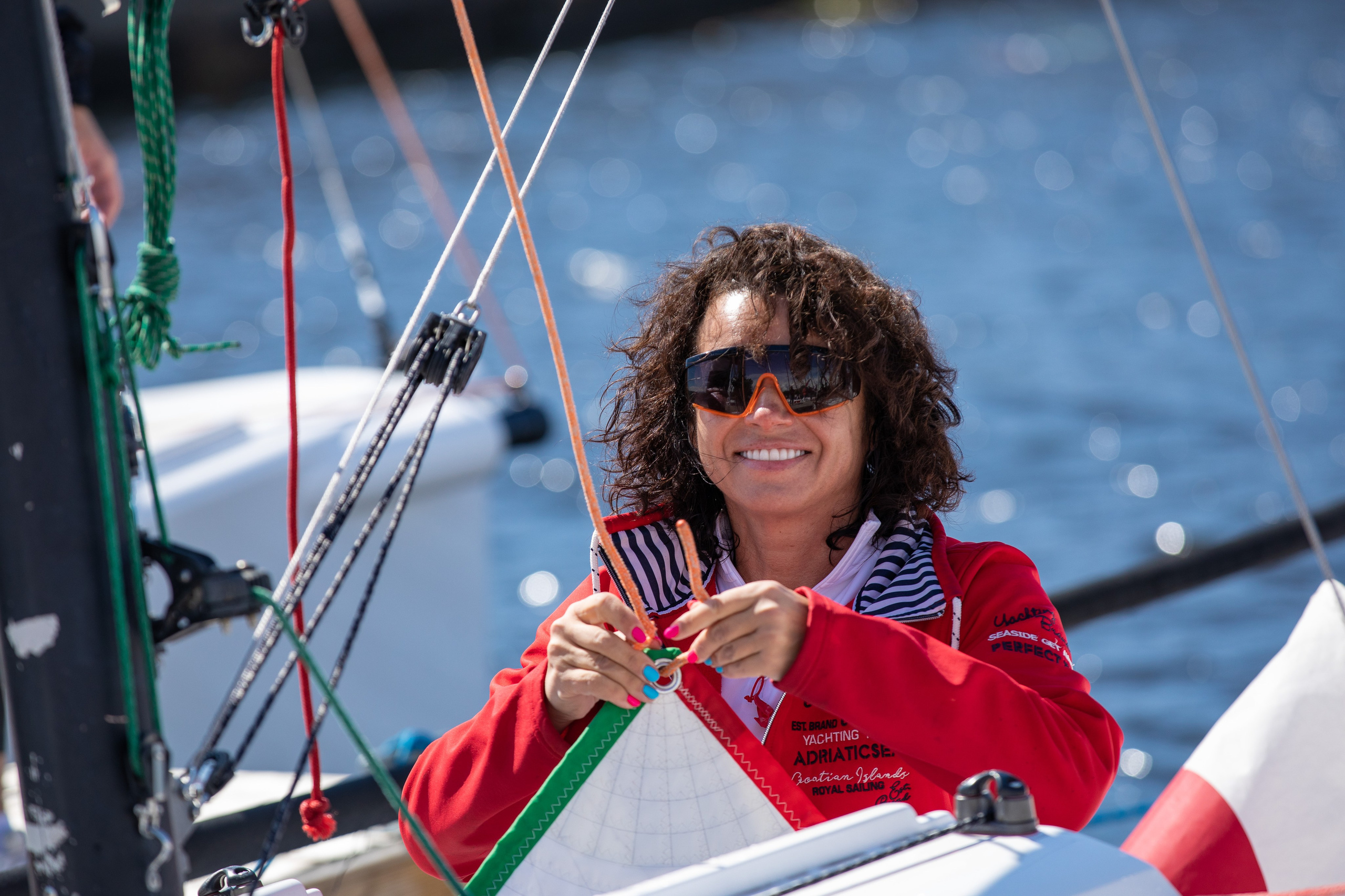 Яхтсменка готовит яхту к выходу на воду. Красивая девушка с кудрявыми волосами и в очках держит парус.