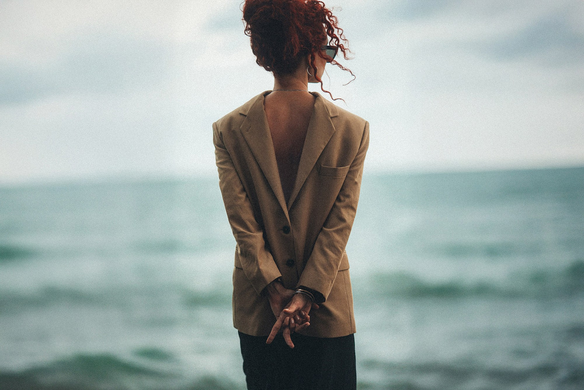 Женщина с ярко-рыжими кудрявыми волосами стоит спиной к камере на фоне бирюзового моря. На ней надет бежевый пиджак задом наперед, обнажающий спину. Руки сцеплены за спиной в замок. Фотограф Галина Скорик.