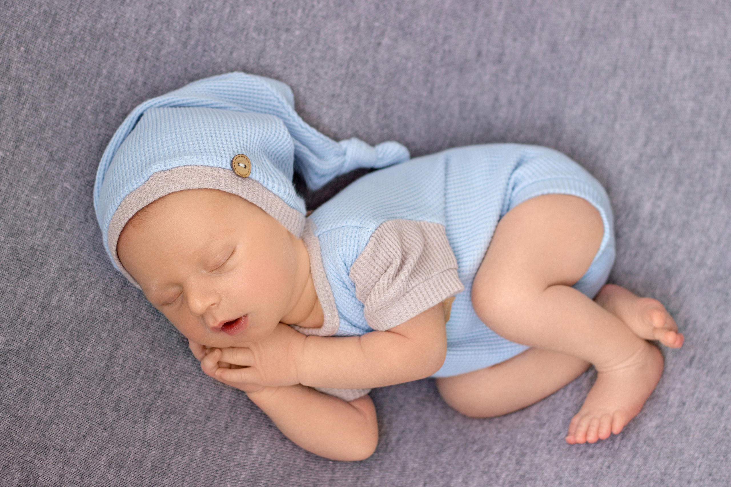 новорожденный мальчик с коликами беспокойным животиком в голубо-сером костюмчике, спит после успокоения, с ручками под щечкой на сером фоне