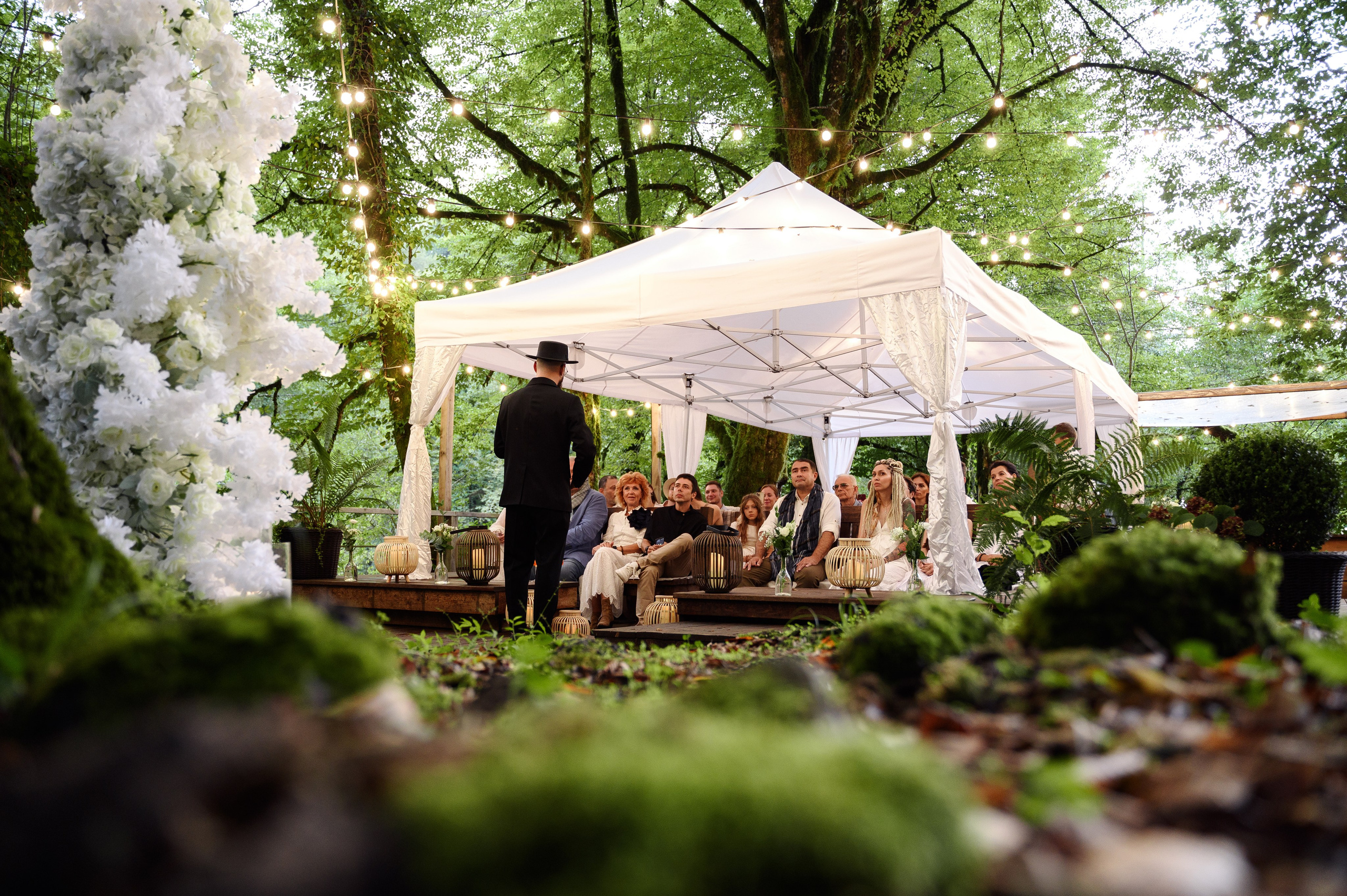 Атмосферный дальний кадр где натянуты шатёр под ним сидят гости и ведущие что-то им говорит все ждут начало свадьбы в Сочи в лесу в глемпинге енот