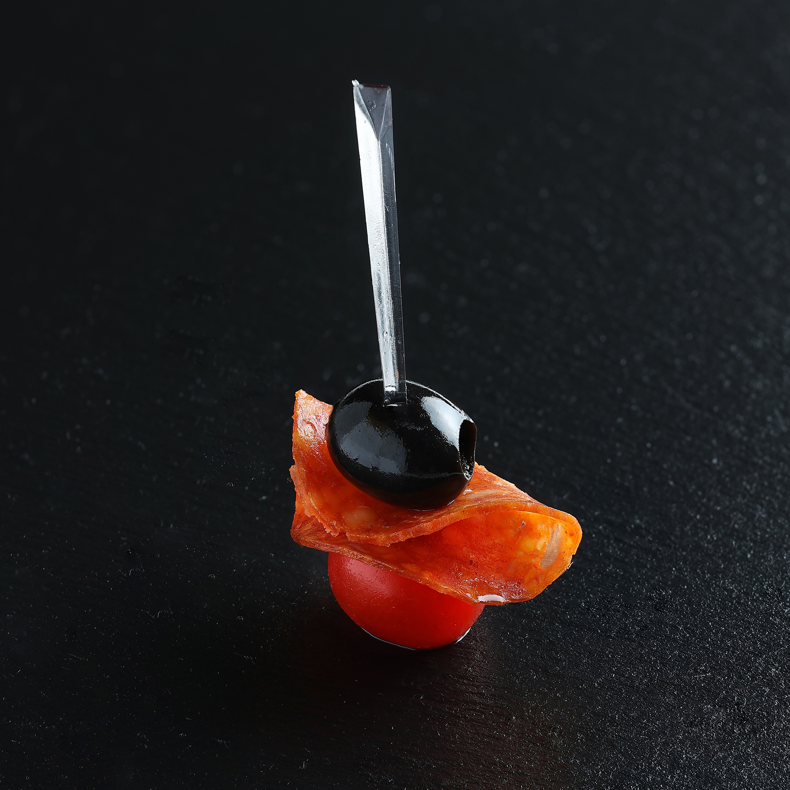 Закуска из помидора черри, салями и чёрной маслины, нанизанная на декоративную шпажку, на тёмной поверхности.