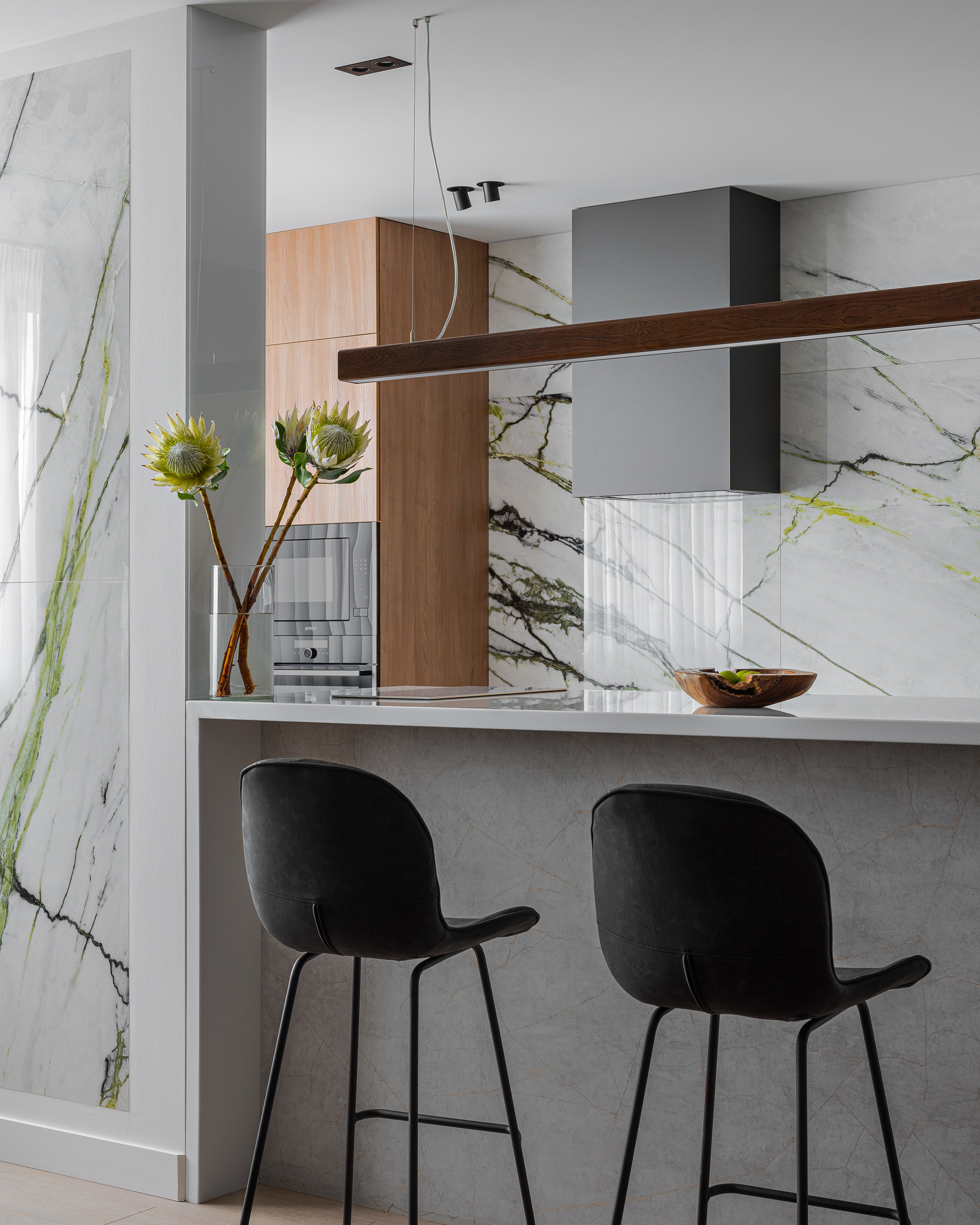 Интерьер кухни с мраморными стенами, белой барной стойкой и черными барными стульями. Декор дополняют минималистичные аксессуары и контраст дерева. Проект дизайнера Алёны Сверловой, фотограф Сергей Денисенко.