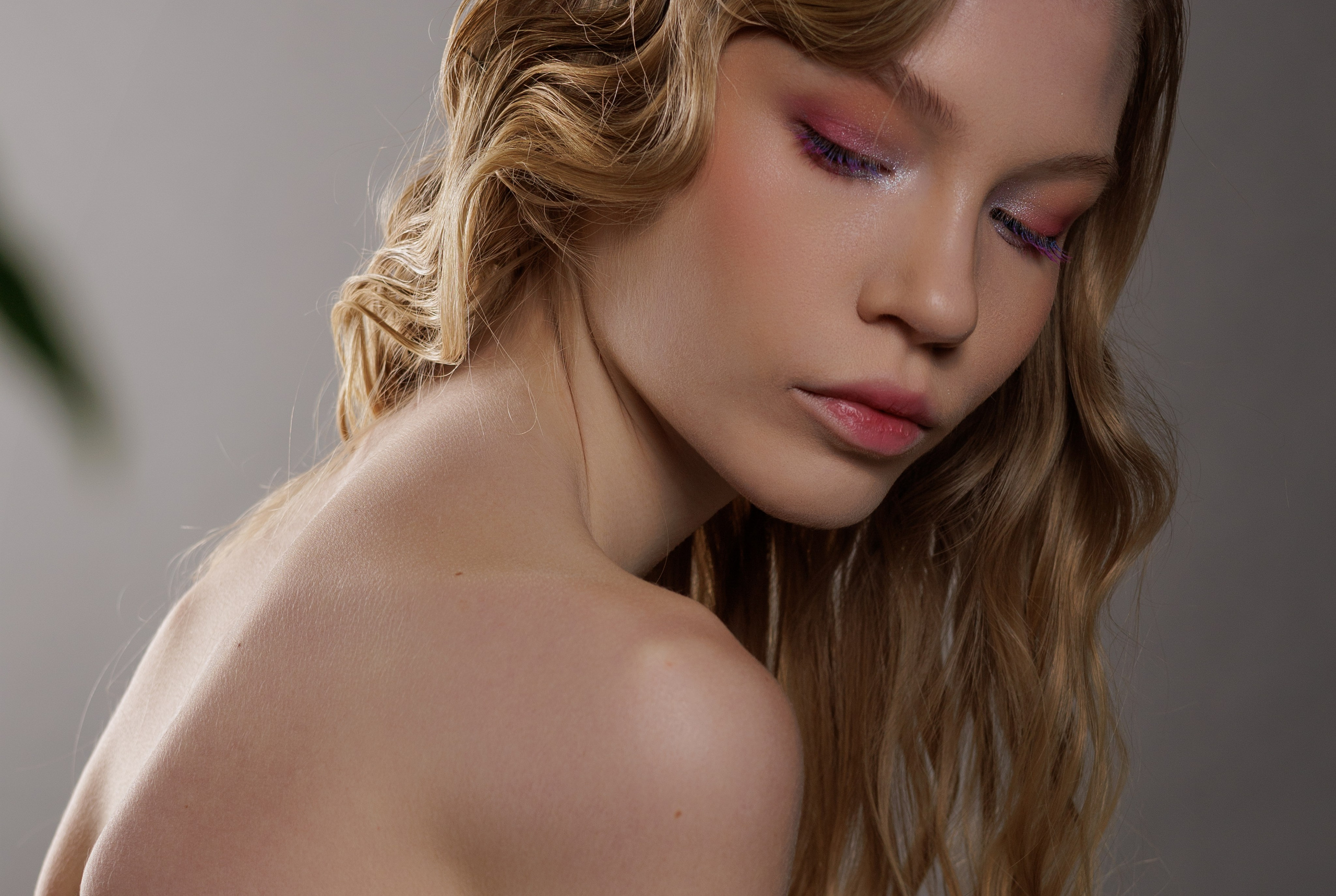 Молодая женщина с волнистыми светлыми волосами и розовым макияжем глаз позирует с закрытыми глазами, передавая спокойное и безмятежное настроение на мягком сером фоне.