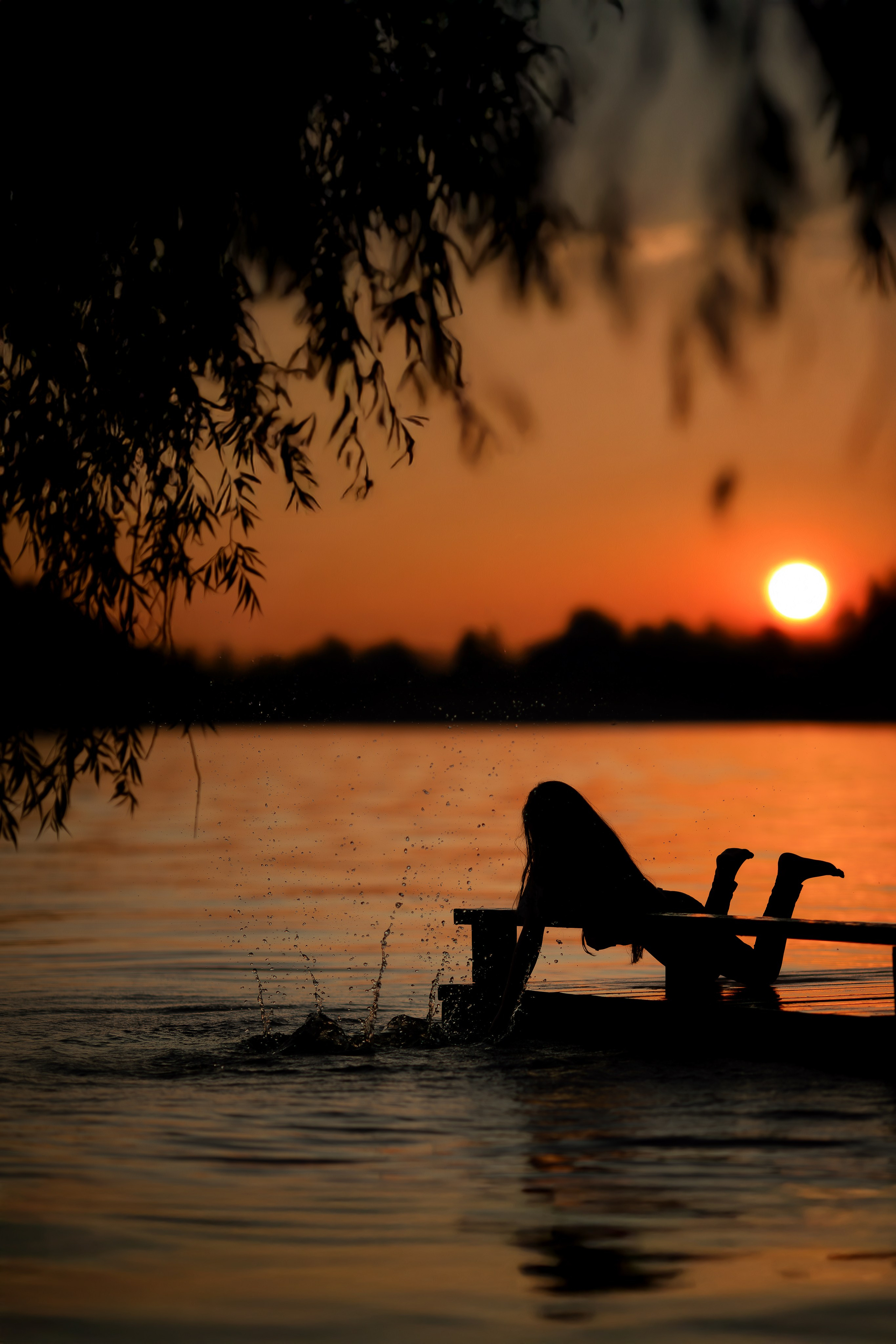 Девочка в силуэте лежит на деревянном мостике у воды, брызги разлетаются в закатном свете, небо и вода окрашены в тёплые оранжевые тона.