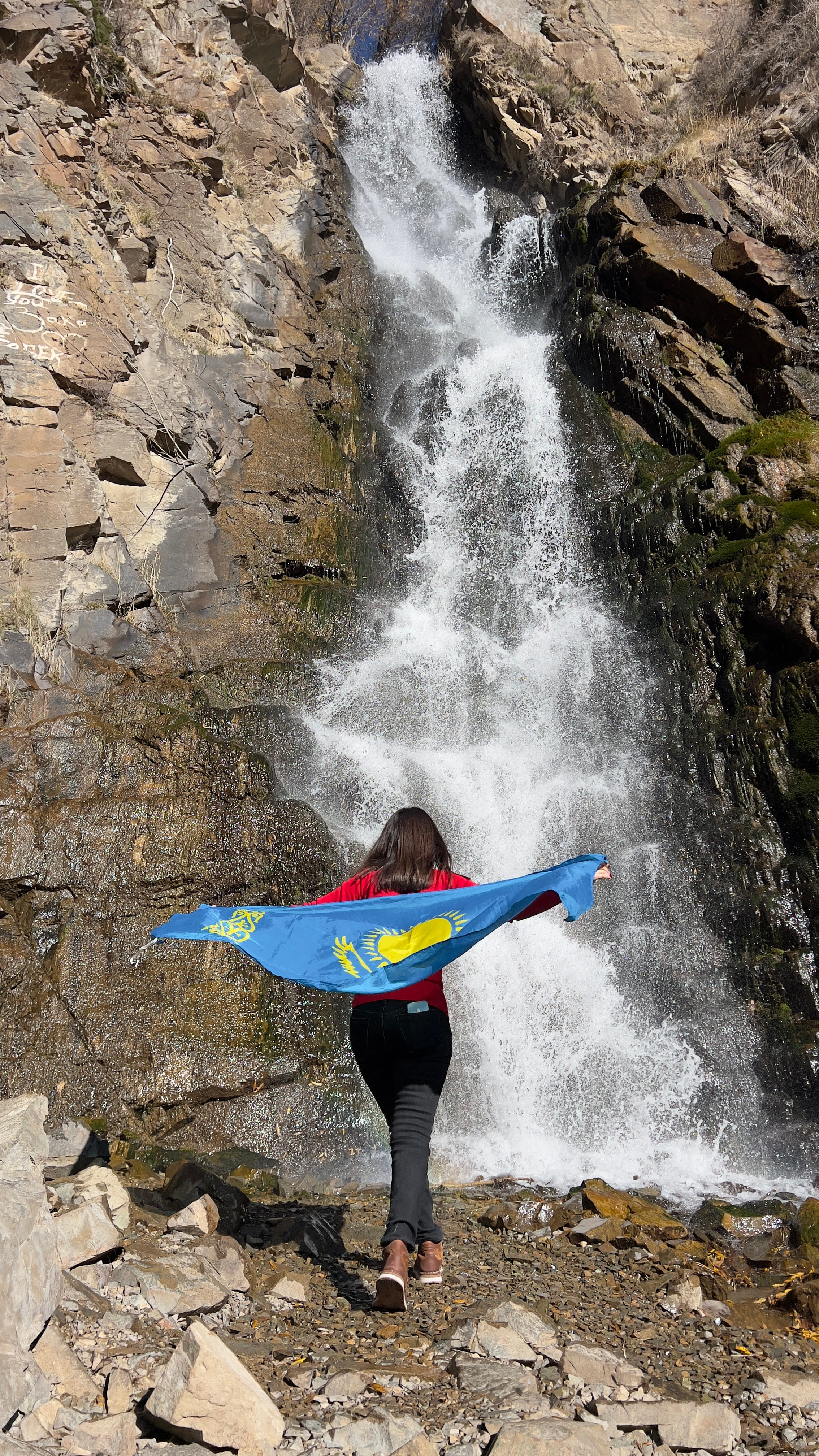 Женщина в красной куртке стоит спиной к камере перед Медвежьим водопадом, раскинув руки с развевающимся флагом Казахстана. На фоне — скалистая местность и бурный поток воды, который ниспадает с высоты. Картина создает ощущение силы природы и гордости за родную страну.
