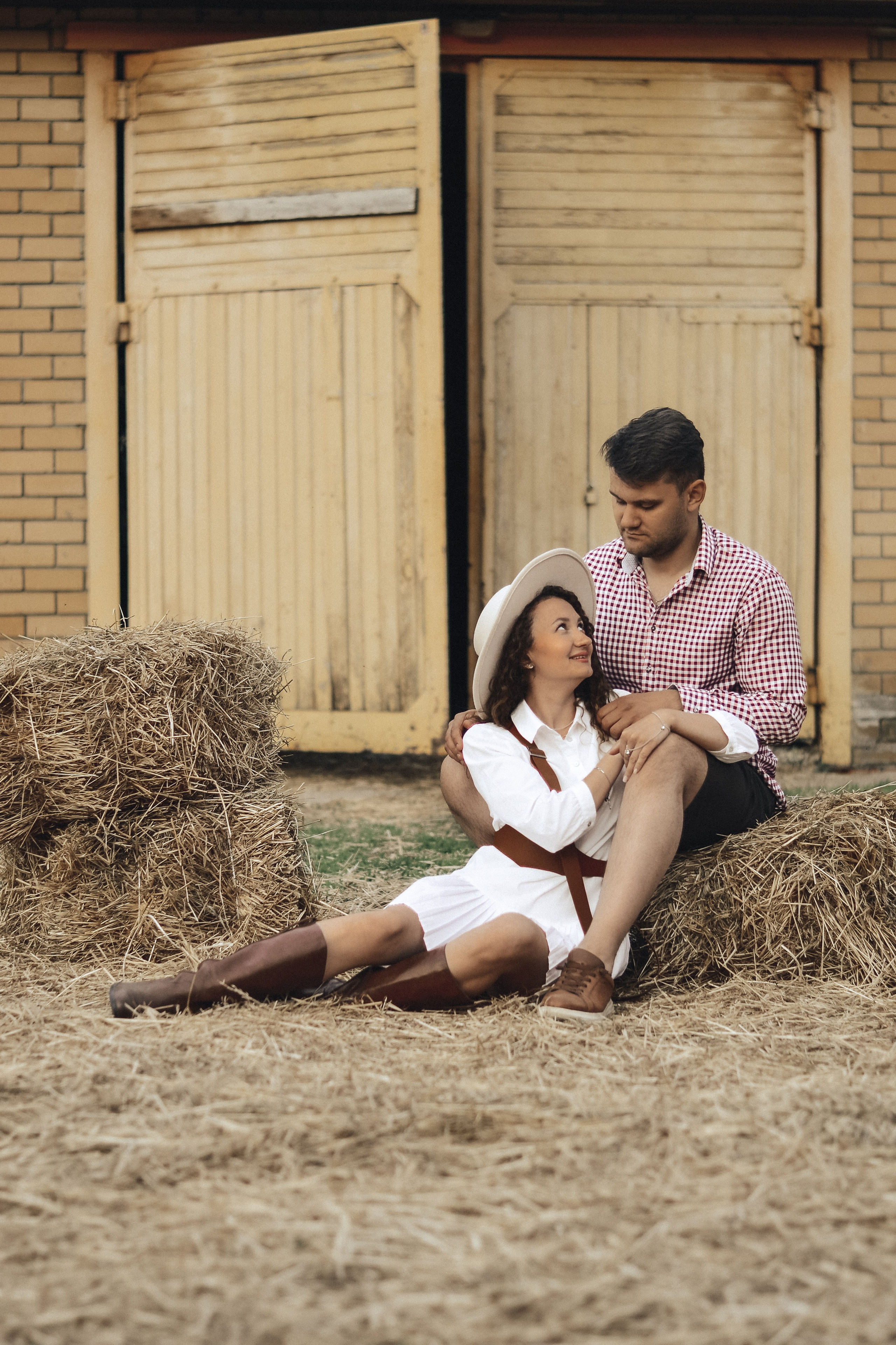 На фотографии изображена пара, которая устроила небольшую фотосессию на фоне старого деревянного амбара. Мужчина одет в клетчатую рубашку и черные шорты, а женщина – в длинное белое платье и большую шляпу. Они сидят на сене, и их позы и взгляды выражают нежность и любовь друг к другу. Элементы деревенского антуража, такие как сено и амбар, добавляют изображению особый колорит и атмосферу уюта.