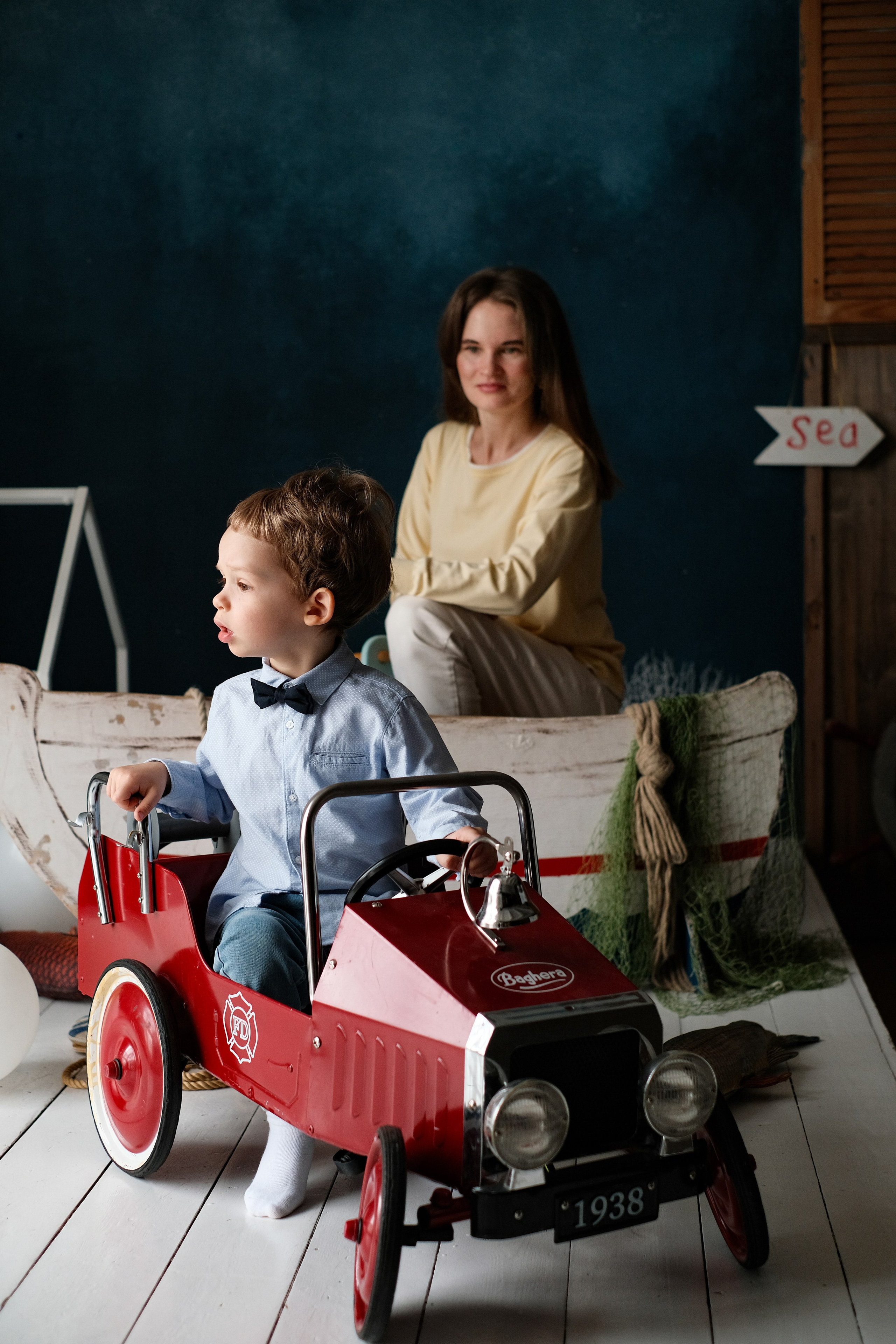 Трехлетний сын сидит в игрушечной машине, на фоне него мама.