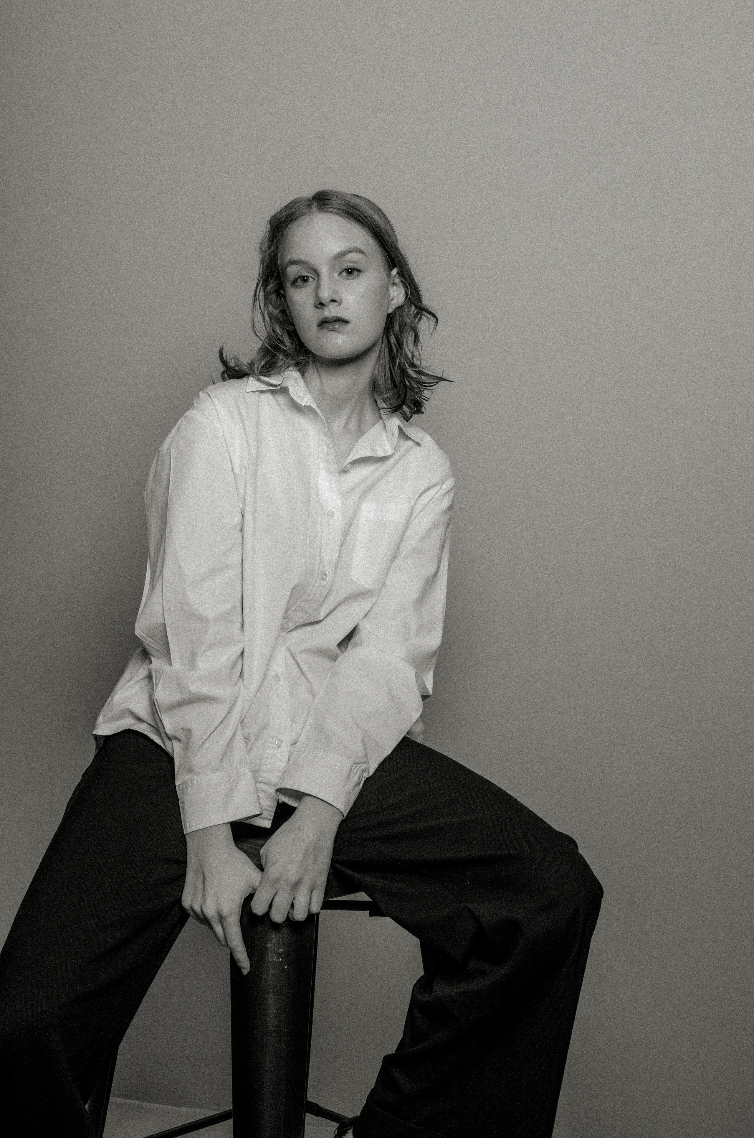 Черно-белая фотография девушки,  сидящей на стуле на простом фоне. Она одета в свободную белую рубашку и темные брюки, излучающие спокойствие и погруженность в себя.