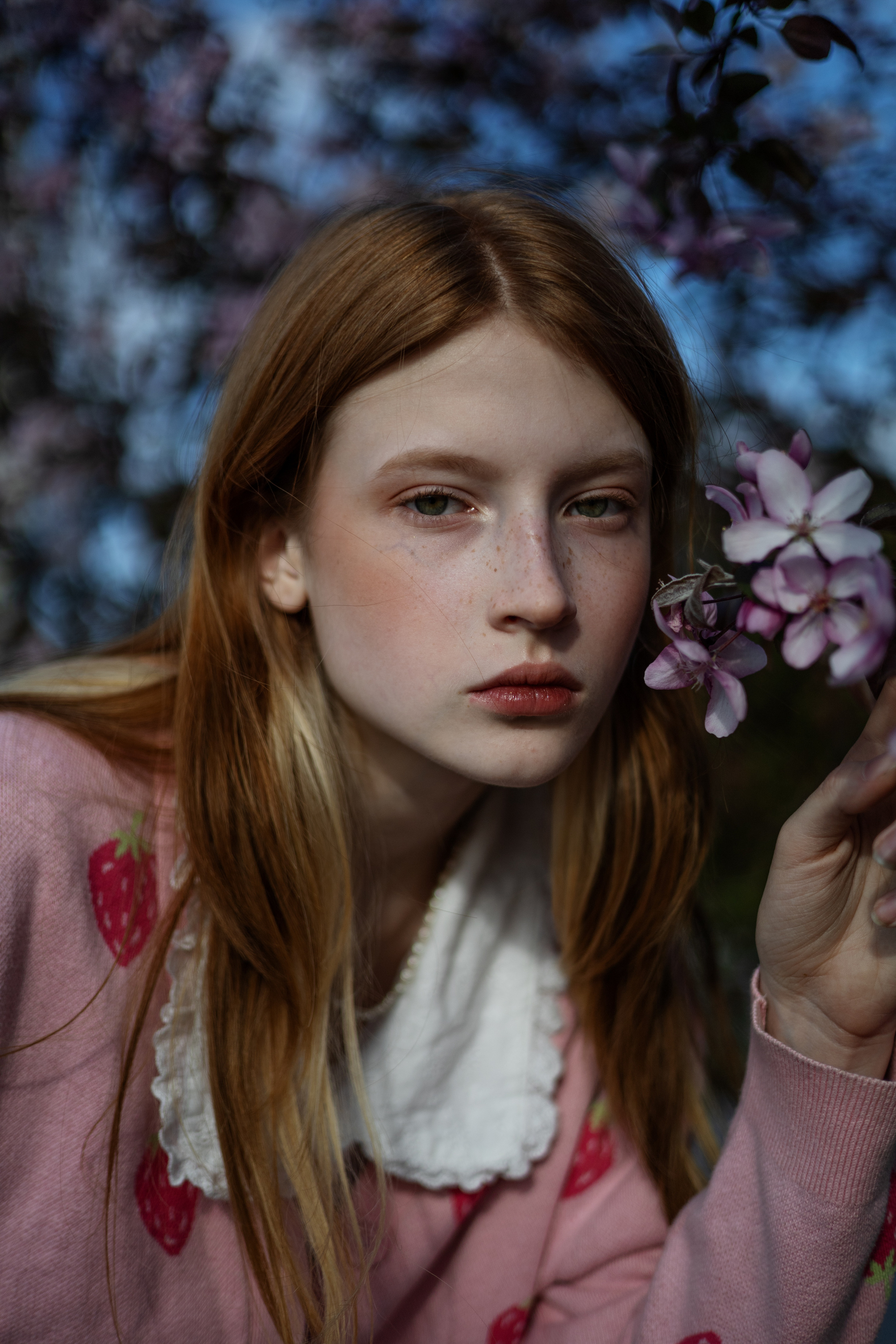 Девушка с длинными рыжими волосами, одетая в розовый свитер с клубникой, держит в руках фиолетовый цветок. Она смотрит в камеру с безмятежным выражением лица.