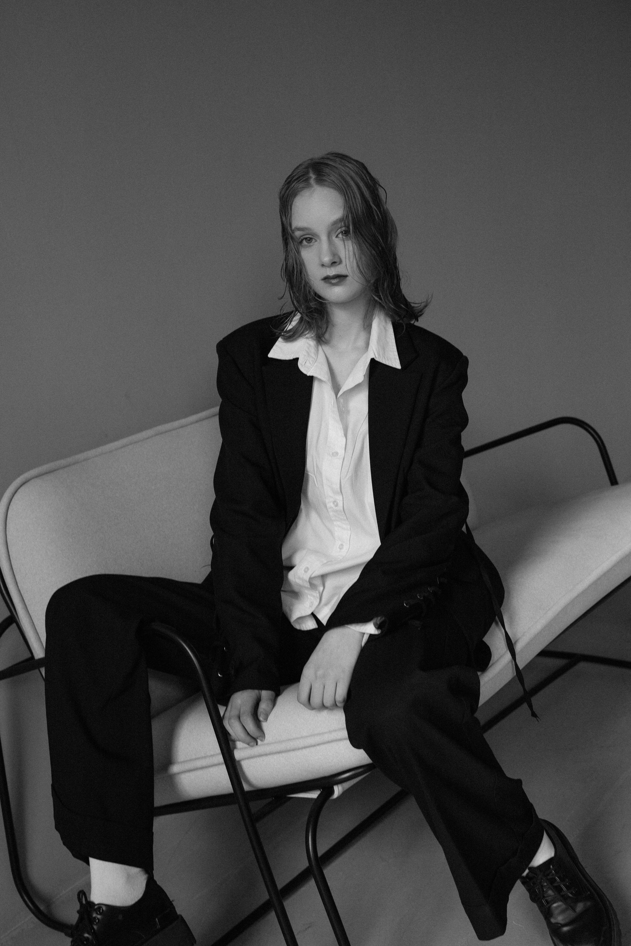Черно-белое изображение  девушки с мокрыми волосами в костюме, сидящей на современном стуле. Непринужденная, уверенная манера  держаться. Обстановка в стиле минимализма.