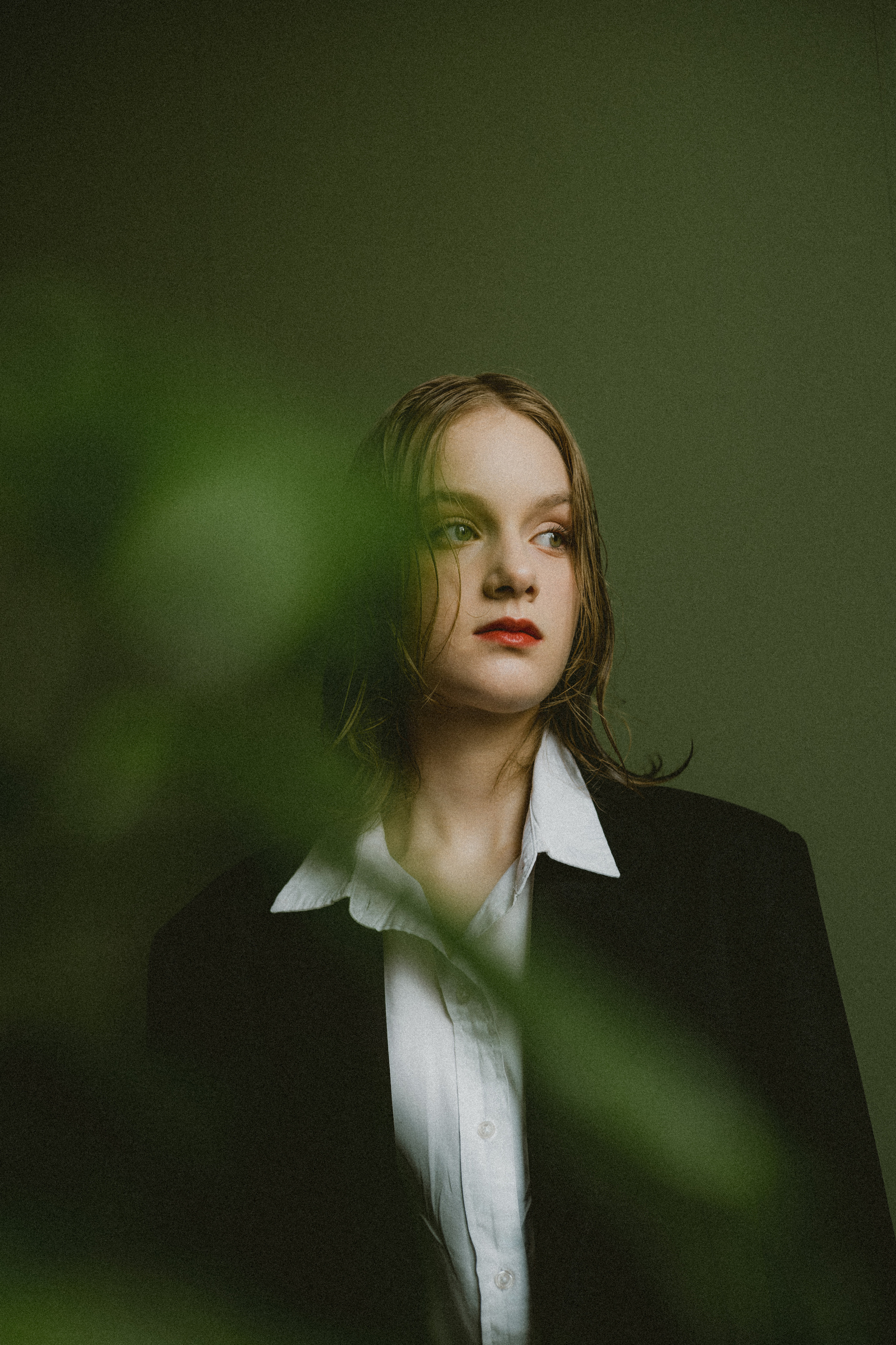 Молодая девушка с мокрыми волосами, одета в черный блейзер и белую рубашку, смотрит за кадром на приглушенно-зеленом фоне, создавая задумчивое настроение.