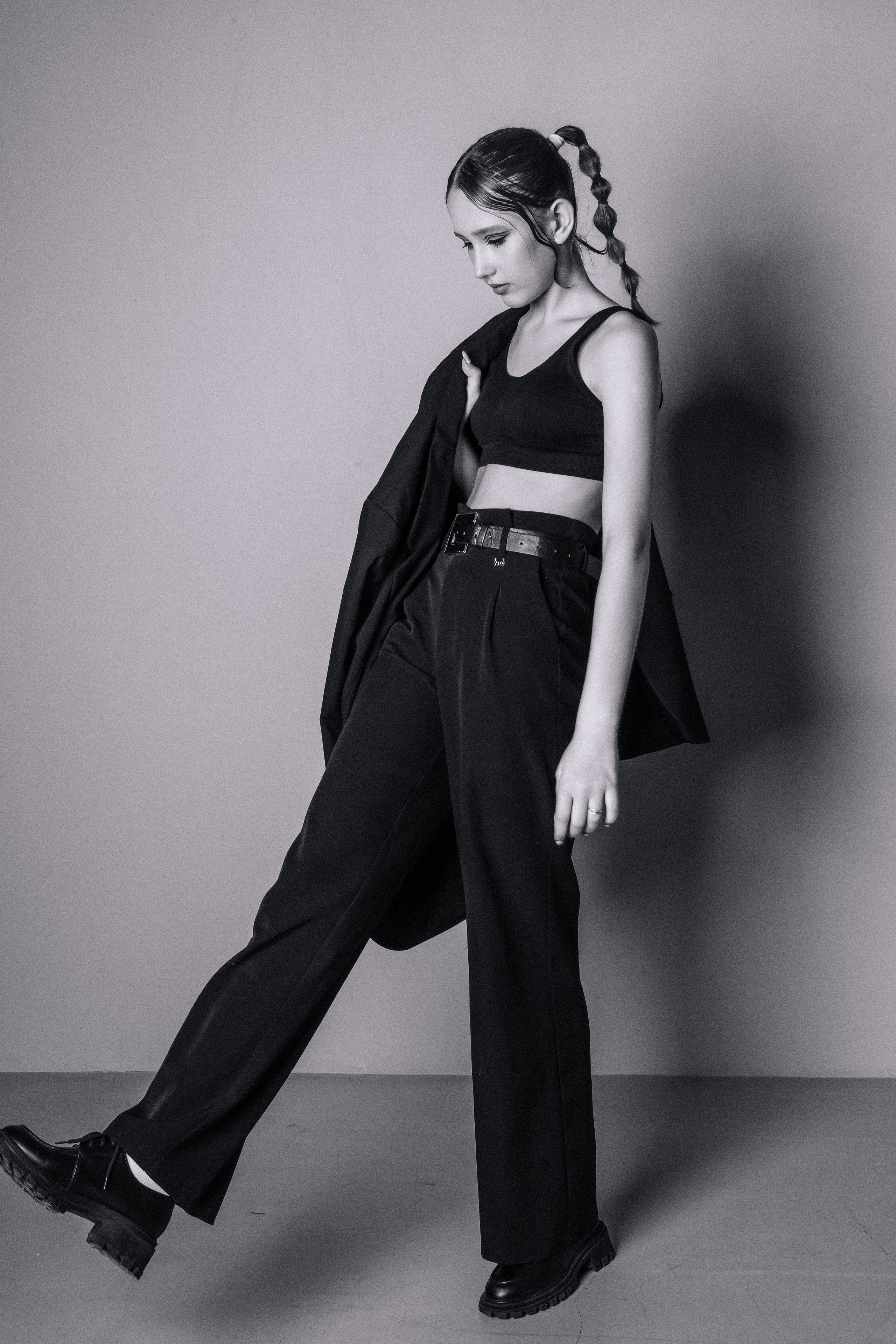 Черно-белая фотография девушки в модном наряде. На ней укороченный топ, брюки на заказ и пальто, перекинутое через плечо, что придает ей уверенный вид. Ее заплетенные в косу волосы и непринужденная поза придают ей стильный вид.