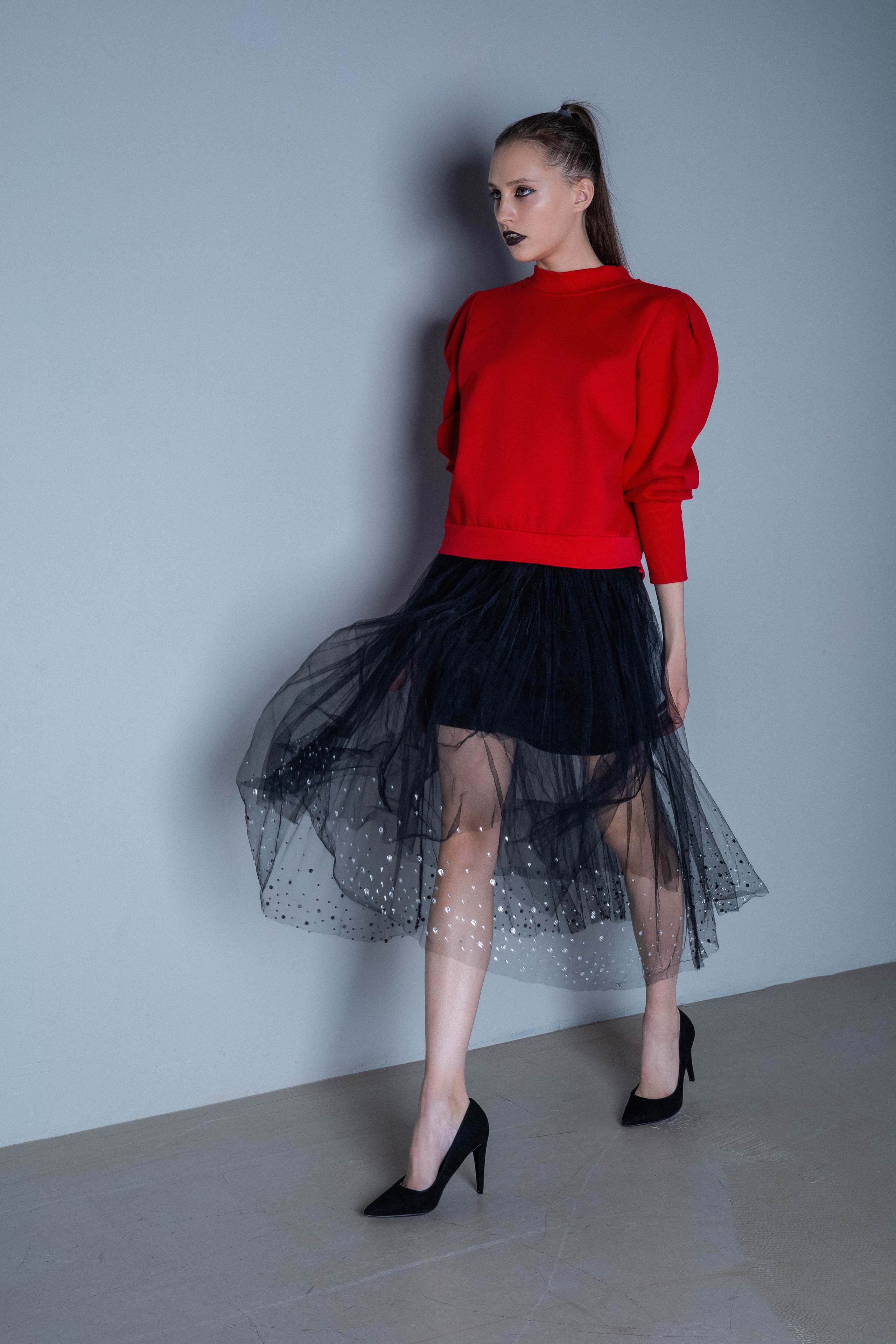 Женщина в ярком красном свитере и прозрачной черной юбке в горошек уверенно позирует. На ней черные туфли на высоких каблуках, создающие стильный контраст.