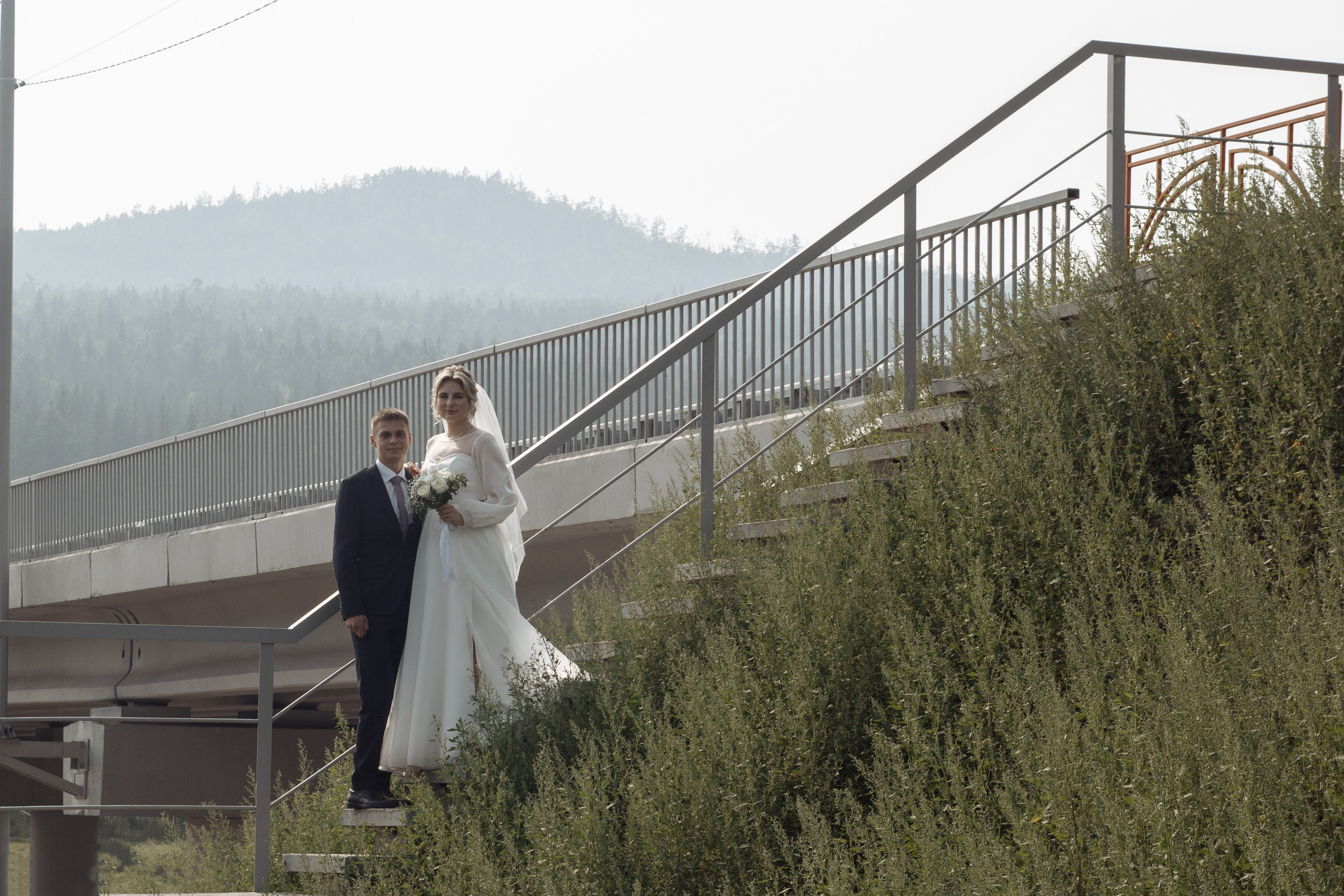 Невеста и жених поднимаются по лестнице на мосту, на заднем плане — лес и горы