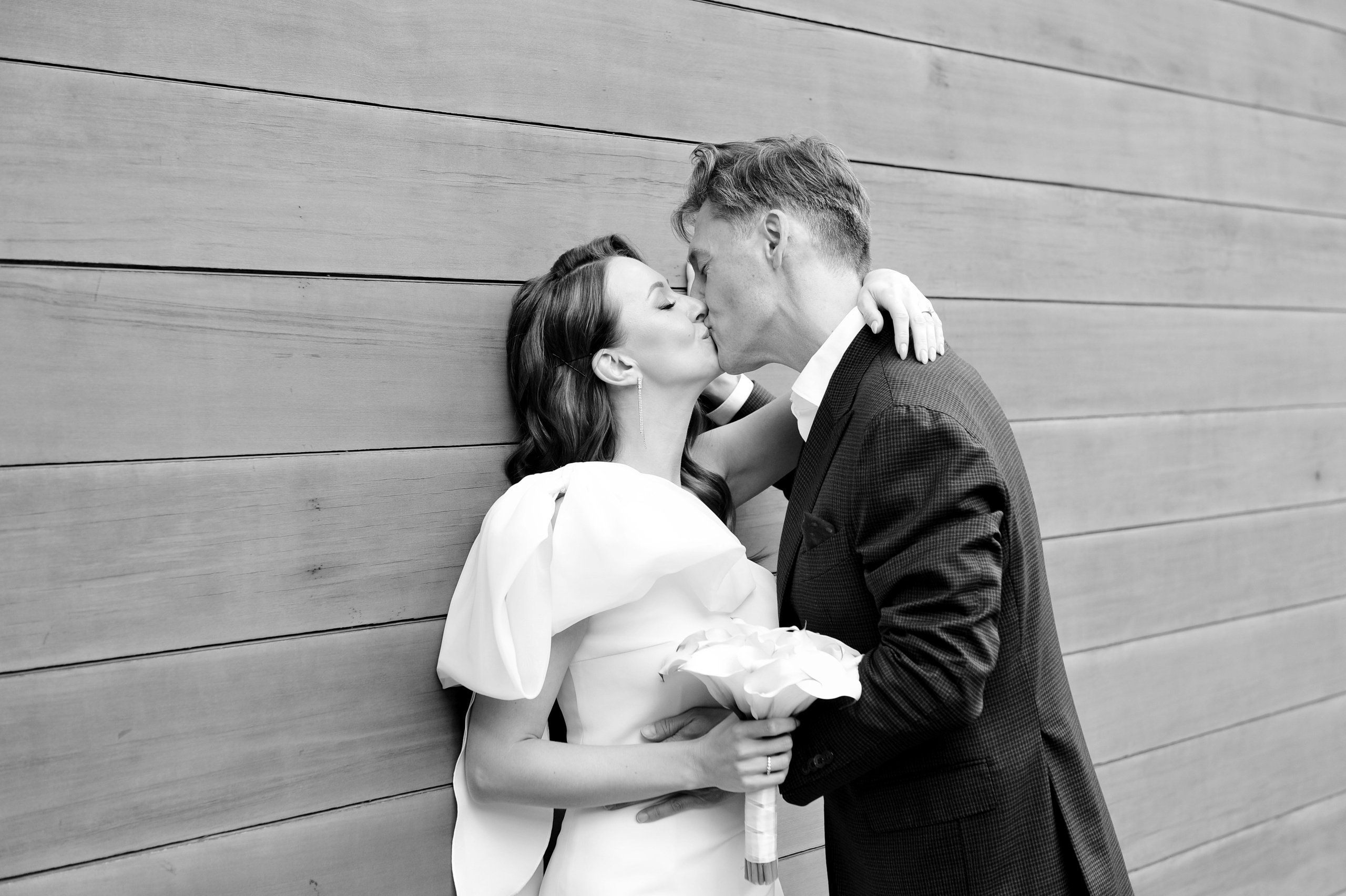 Свадьба в ЗАГСе Барвиха Luxury Village летом, жених и невеста стоят у деревянной стены здания, поцелуй, чёрно-белая фотография