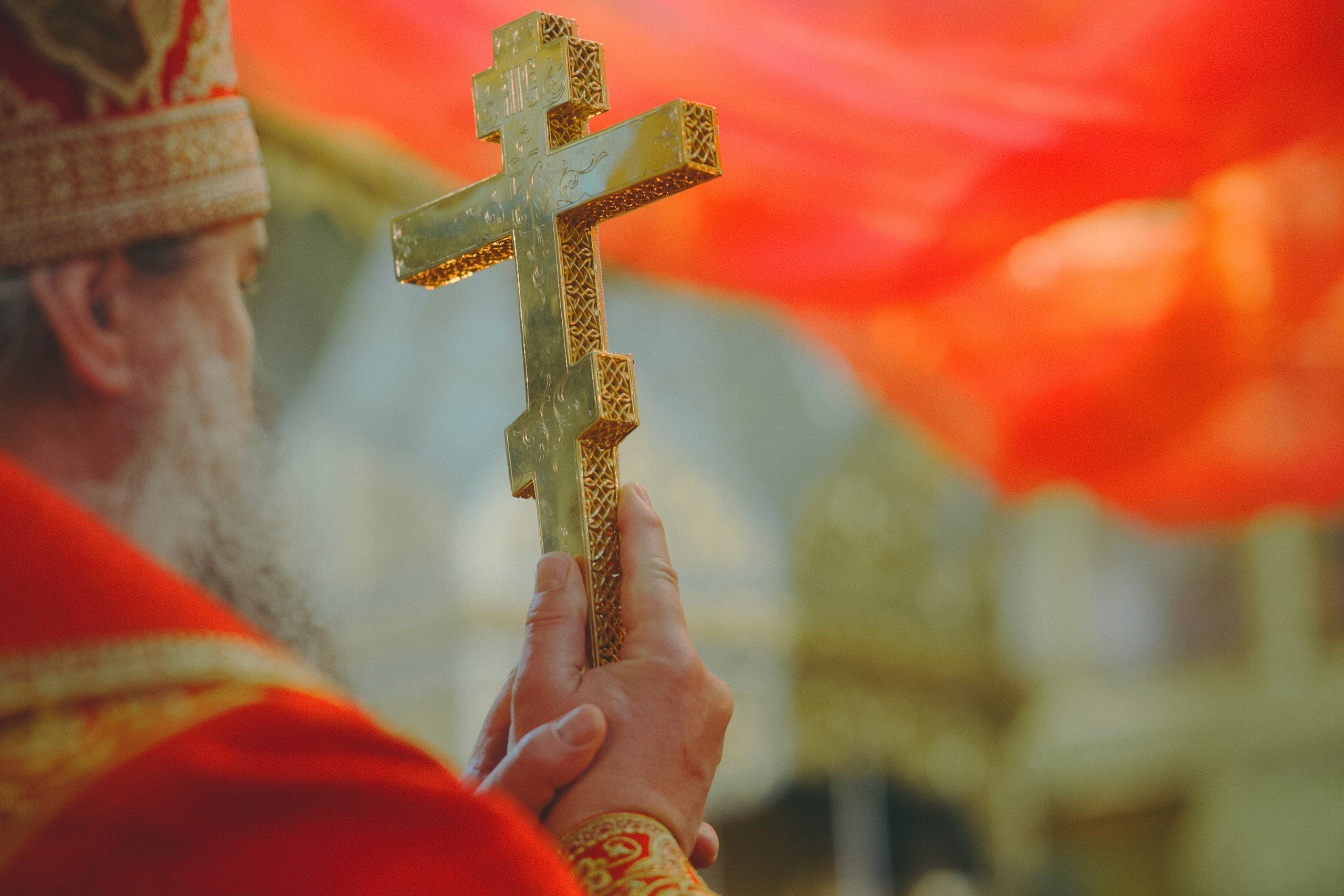 Крупный план: священник в красной фелони держит в руках золотой напрестольный крест с орнаментом. Фон — красный покров.