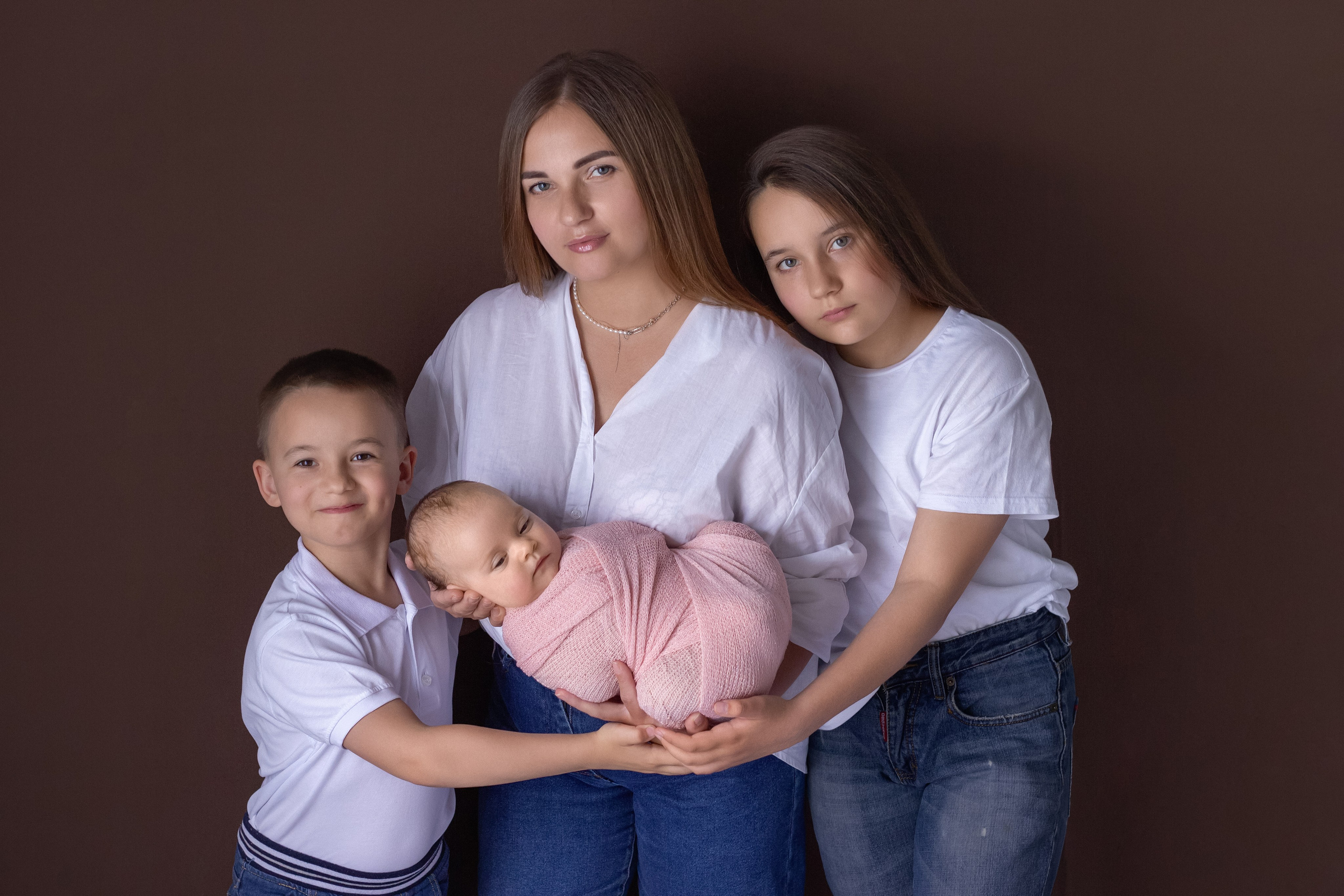портрет счастливой семьи - мамы, дочери, сына и младшей новорожденной, все одеты в белые футболки и синие джинсы, все обнимаются